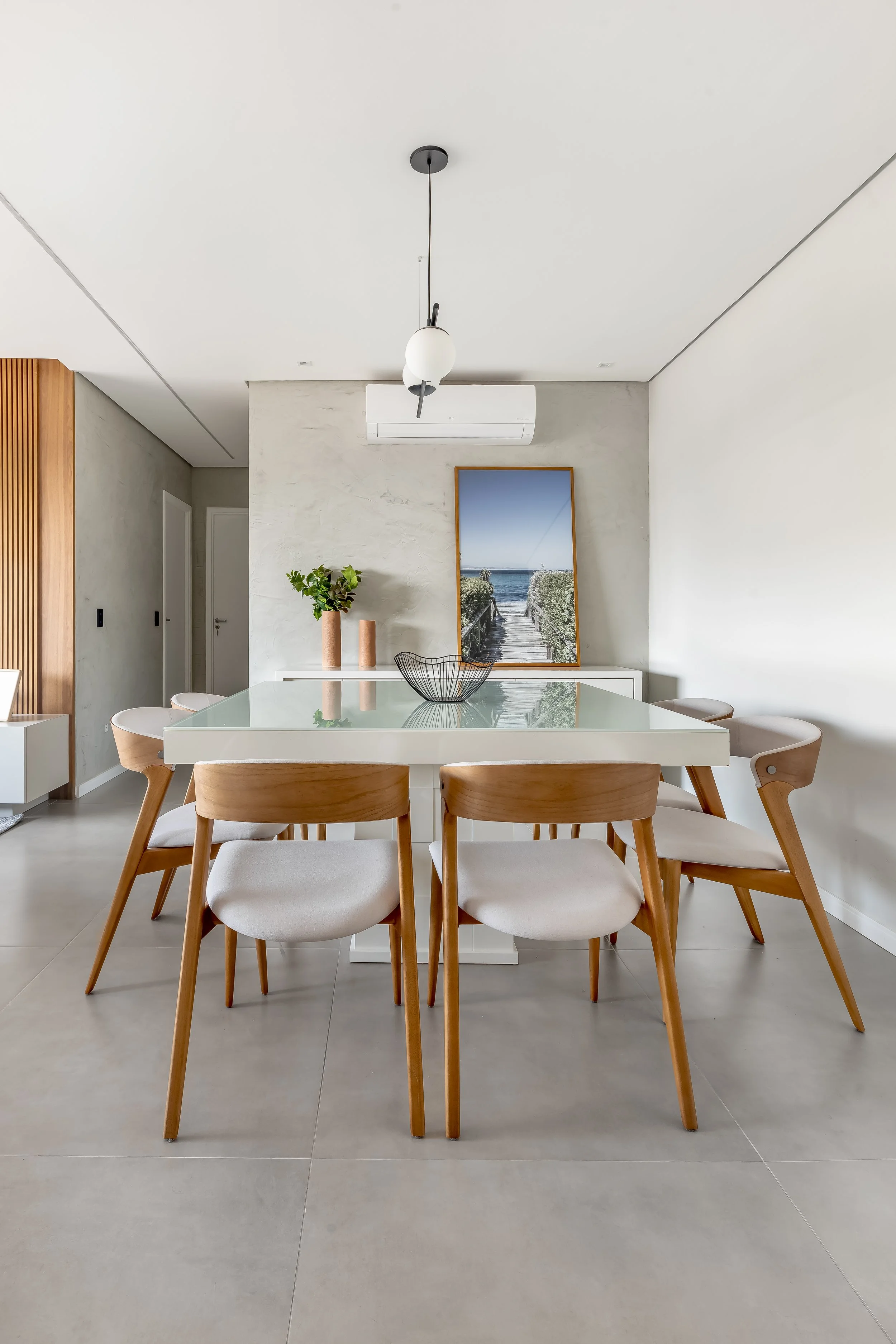 Sala de jantar moderna com mesa de vidro branca, seis cadeiras de madeira com assento branco, decoração minimalista com quadro de fotografia de uma passagem para a praia e dois vasos com plantas ao fundo.