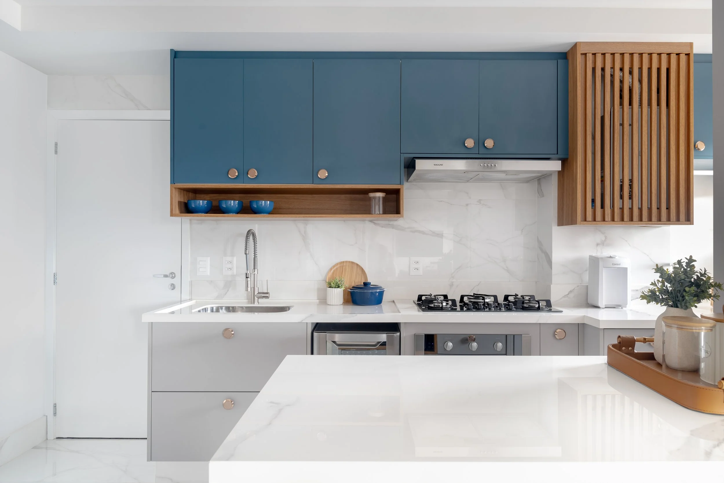 Cozinha moderna com armários azuis, detalhes de madeira, bancada de mármore branco, pia e cooktop de gás, decorada com vasos verdes e utensílios