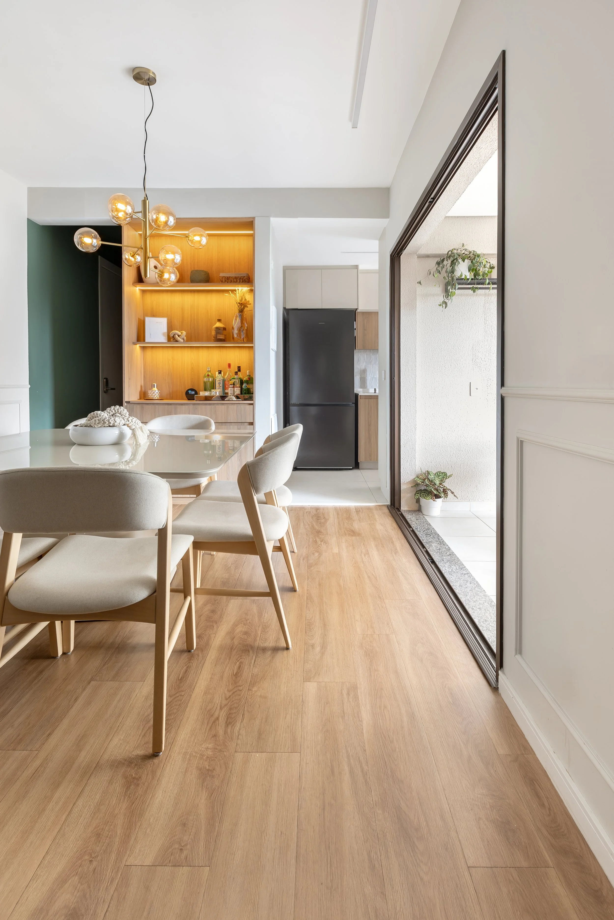 Sala de jantar com mesa de madeira clara, cadeiras brancas e uma luminária moderna com lâmpadas de vidro. Ao fundo, uma parede com estantes de madeira iluminadas e uma geladeira preta. A porta de correr leva para uma varanda com plantas.