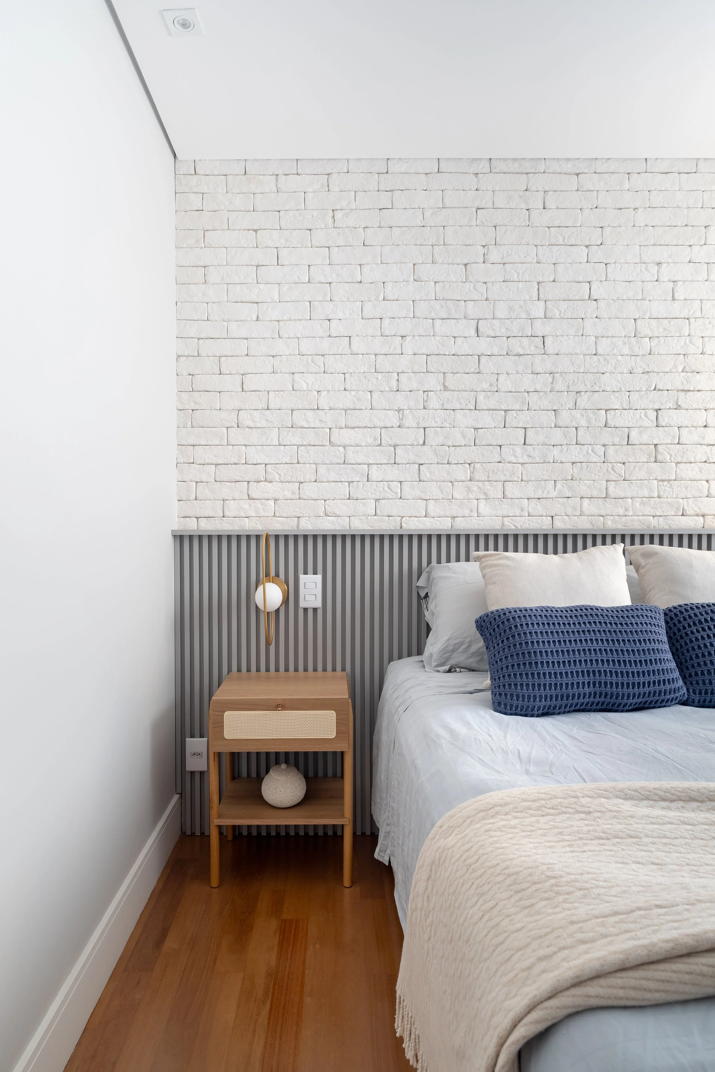 Quarto com parede de tijolos brancos, cabeceira cinza, cama com travesseiros brancos e azuis, cômoda de madeira ao lado com objeto decorativo de bola de pedra e lâmpada de parede com design moderno.
