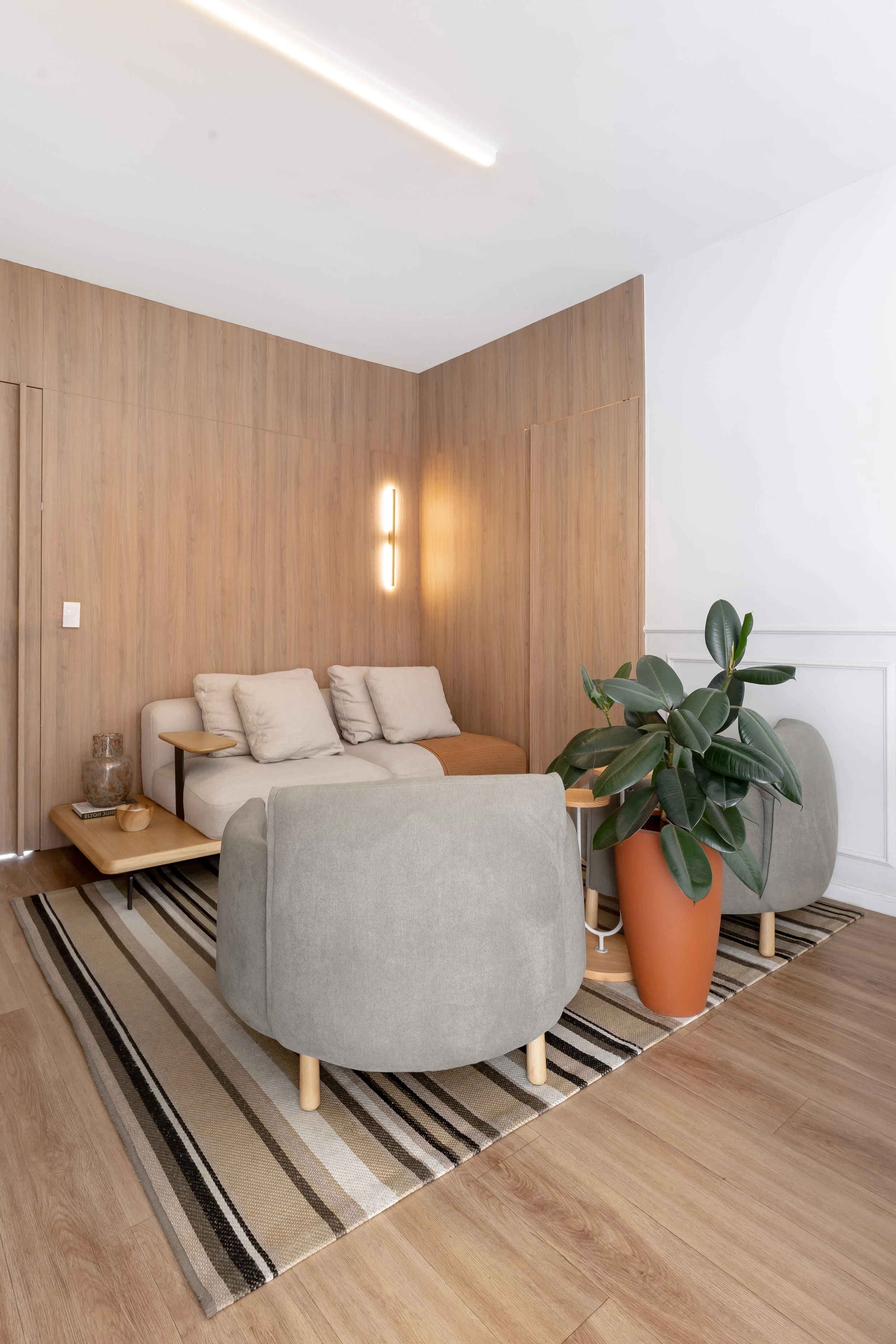 Sala de estar com parede de madeira e iluminação suave, sofá bege com almofadas, tapete listrado, mesa de centro, vaso grande com planta verde no canto.