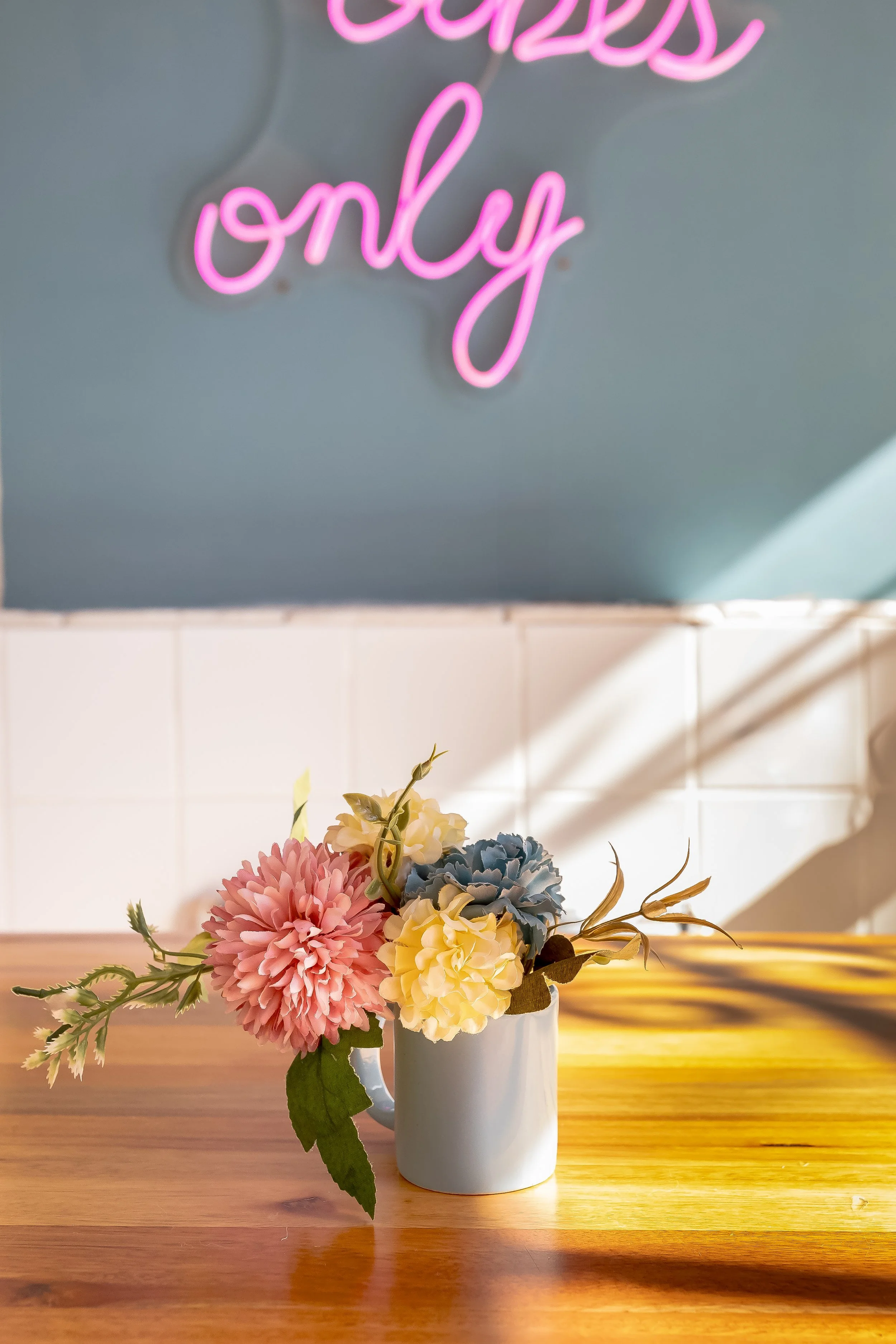 Vaso com flores artificiais rosa, amarelo, azul e verde, sobre mesa de madeira, com mural de neon rosa com escrita em inglês no fundo.