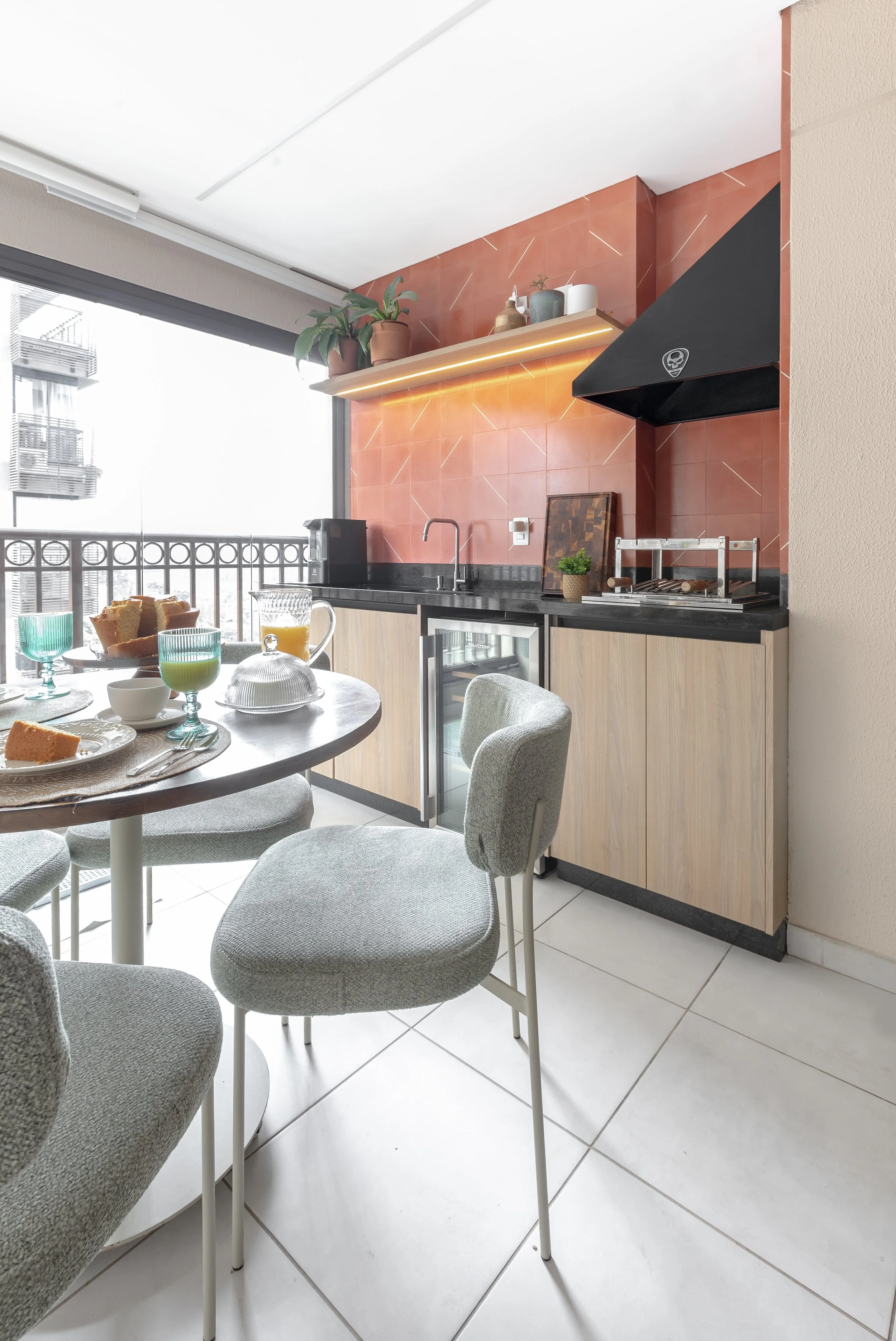 Sala de jantar com mesa redonda, cadeiras confortáveis, uma sala de cozinha ao fundo com armários em madeira clara, bancada em granito preto, uma parede com azulejos vermelhos, uma prateleira com plantas e objetos decorativos, janela grande com varan