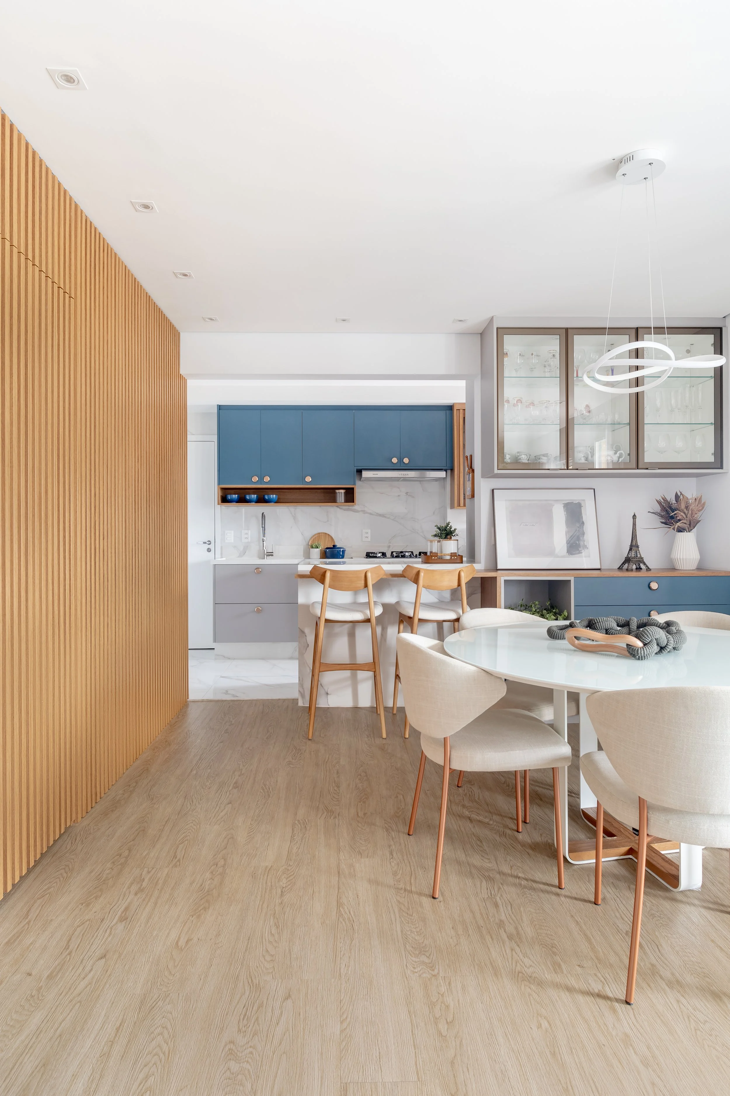 Cozinha moderna com armários azuis, bancada de mármore, parede com vidro de vidro para exibir louças, mesa de jantar com cadeiras creme e uma parede de madeira na esquerda.
