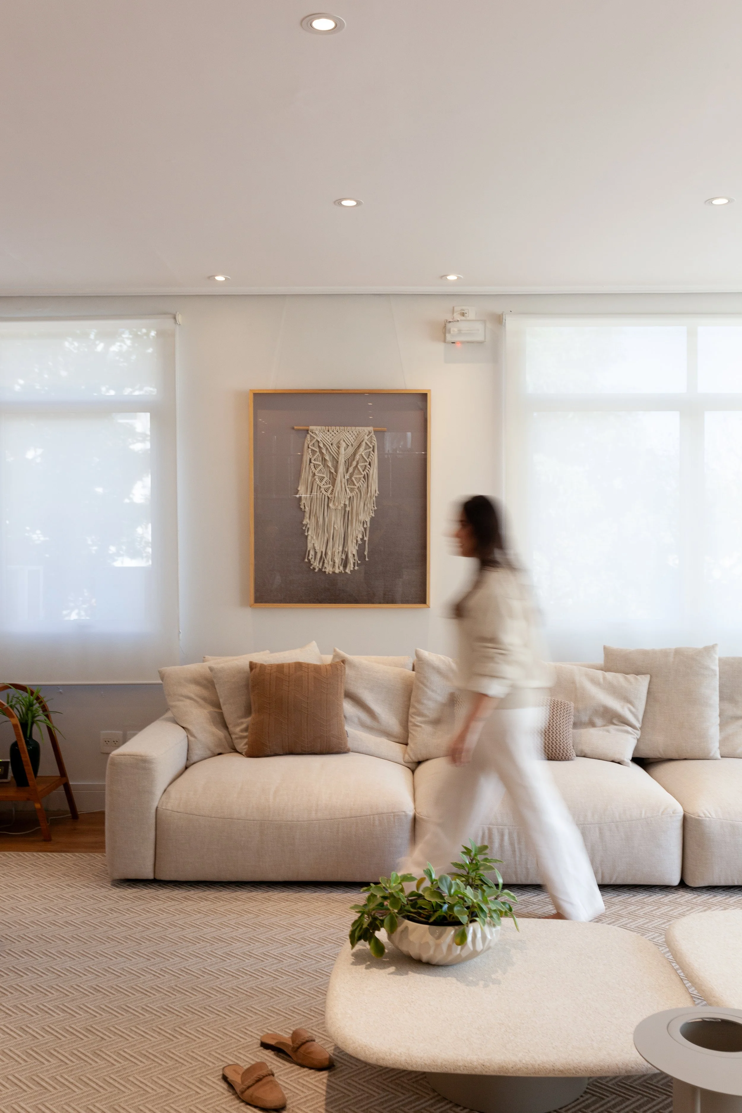 Sala de estar com sofá bege, almofadas, puf de canto, plantas e quadro decorativo na parede, com uma pessoa caminhando.