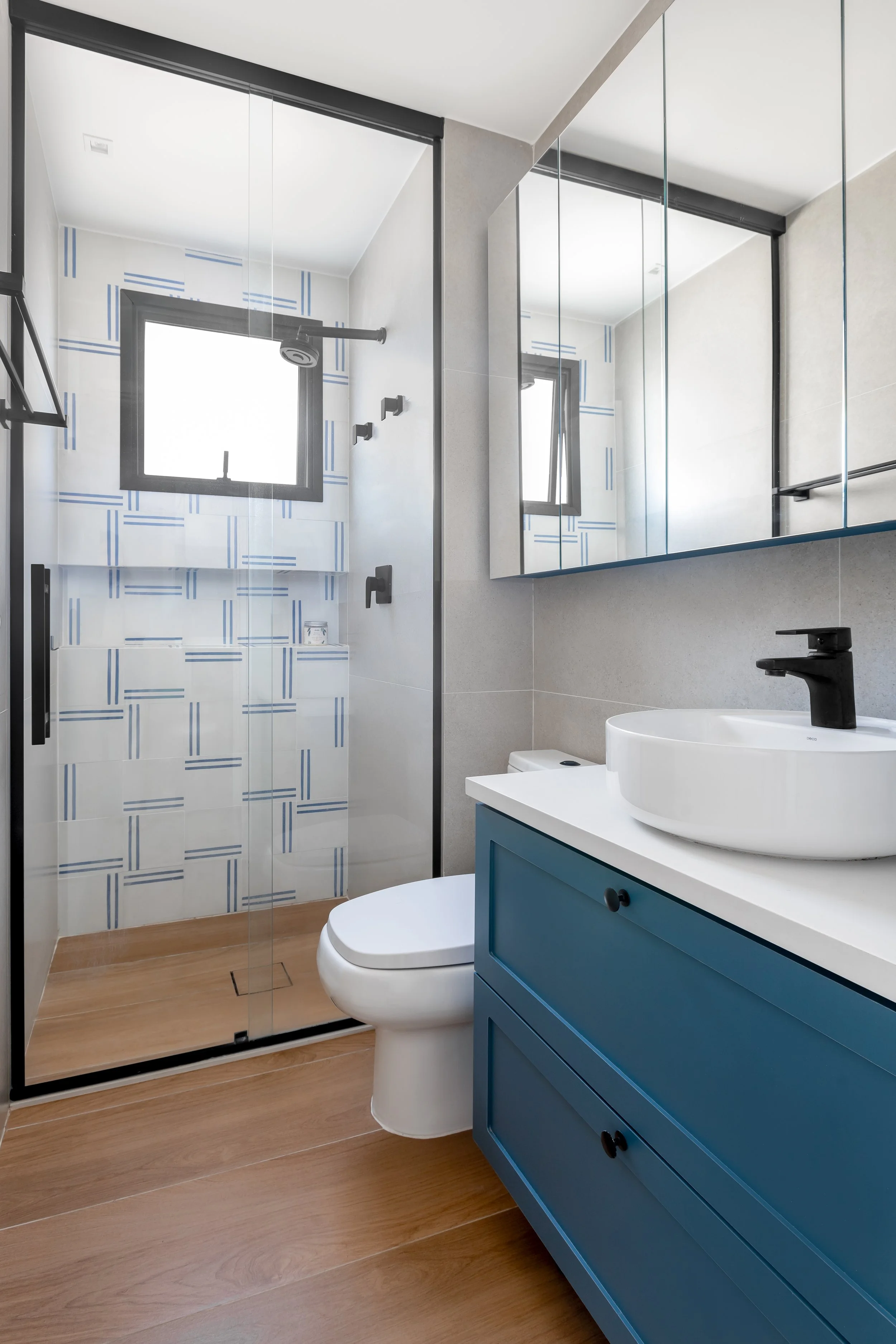 Banheiro moderno com azulejos azuis e brancos na área do chuveiro, ducha com janelas pequenas e espelhos, pia branca em móvel azul, vaso sanitário branco, piso de madeira, acabamento clean e iluminação natural.