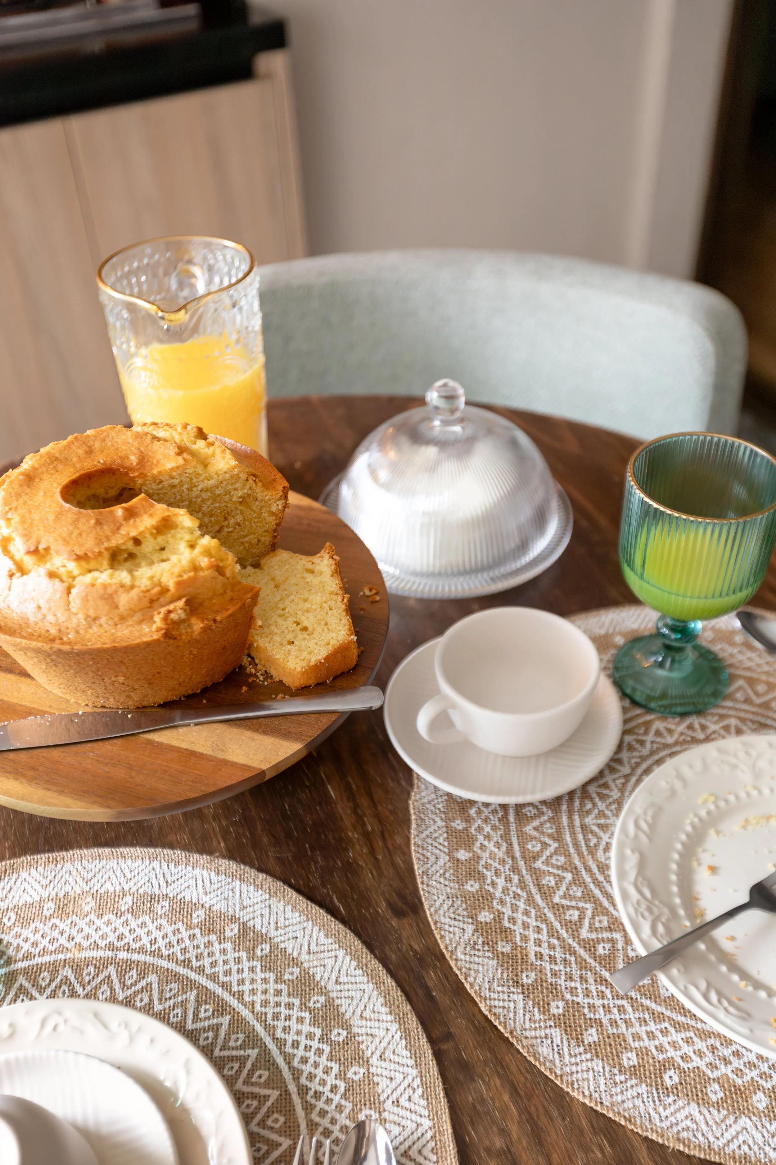 Mesa de café da manhã com bolo de fubá, suco de laranja, copo de vidro colorido, xícara branca, prato com manteiga, colher, garfo e faca, tudo sobre uma toalha de renda.