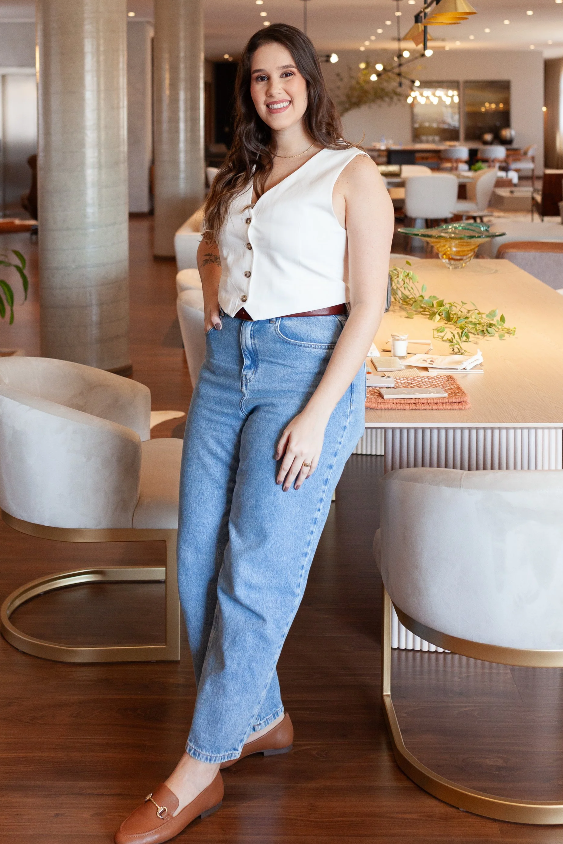 Mulher sorridente com cabelo castanho escuro, vestindo uma blusa branca de botões, blusa sem mangas, jeans azul claro, sapatos marrom e uma pulseira, posando em um ambiente interno de estilo moderno e aconchegante.