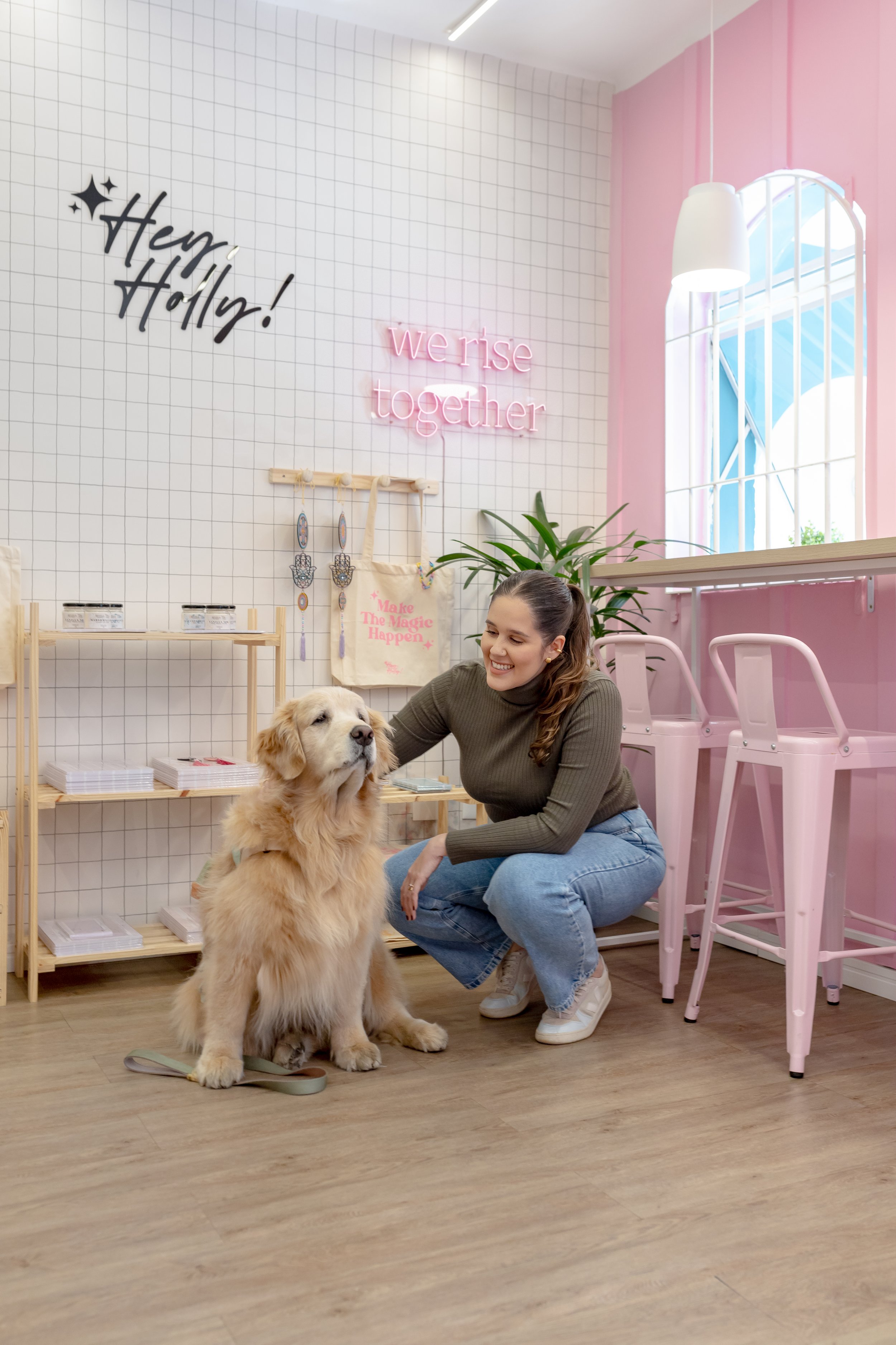 Mulher sorridente brincando com um cachorro golden retriever dentro de um espaço decorado com paredes brancas, rosa e um letreiro de neon rosa que diz "we rise together", e uma parede com palavras "Hey, Holly!" e uma tábua com objetos decorativos.