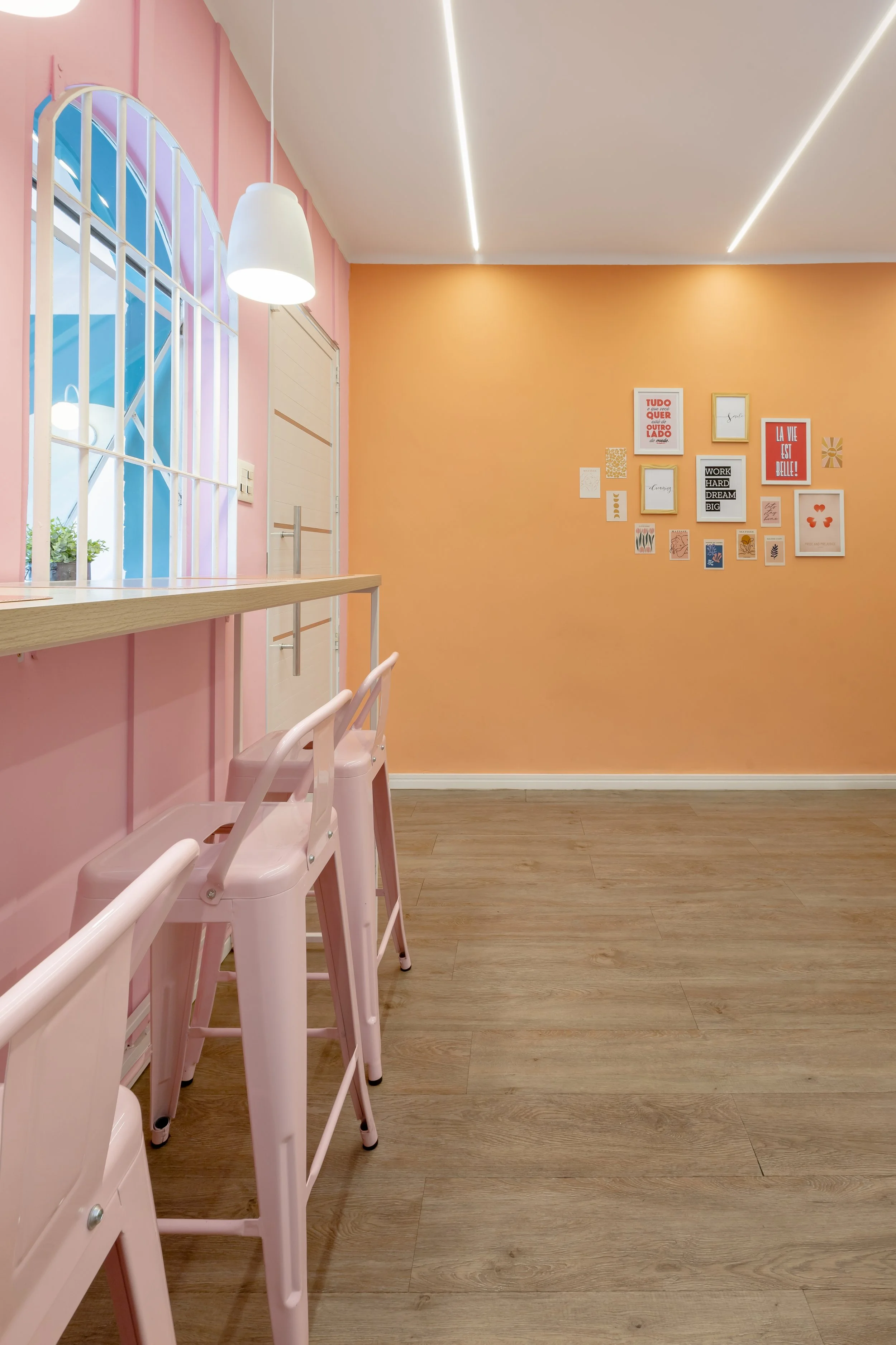 Interior de um espaço decorado com paredes em cores pastel, incluindo uma parede laranja e uma parede rosa com uma janela com grades brancas, cadeiras altas rosa, piso de madeira e uma parede com quadros decorativos.
