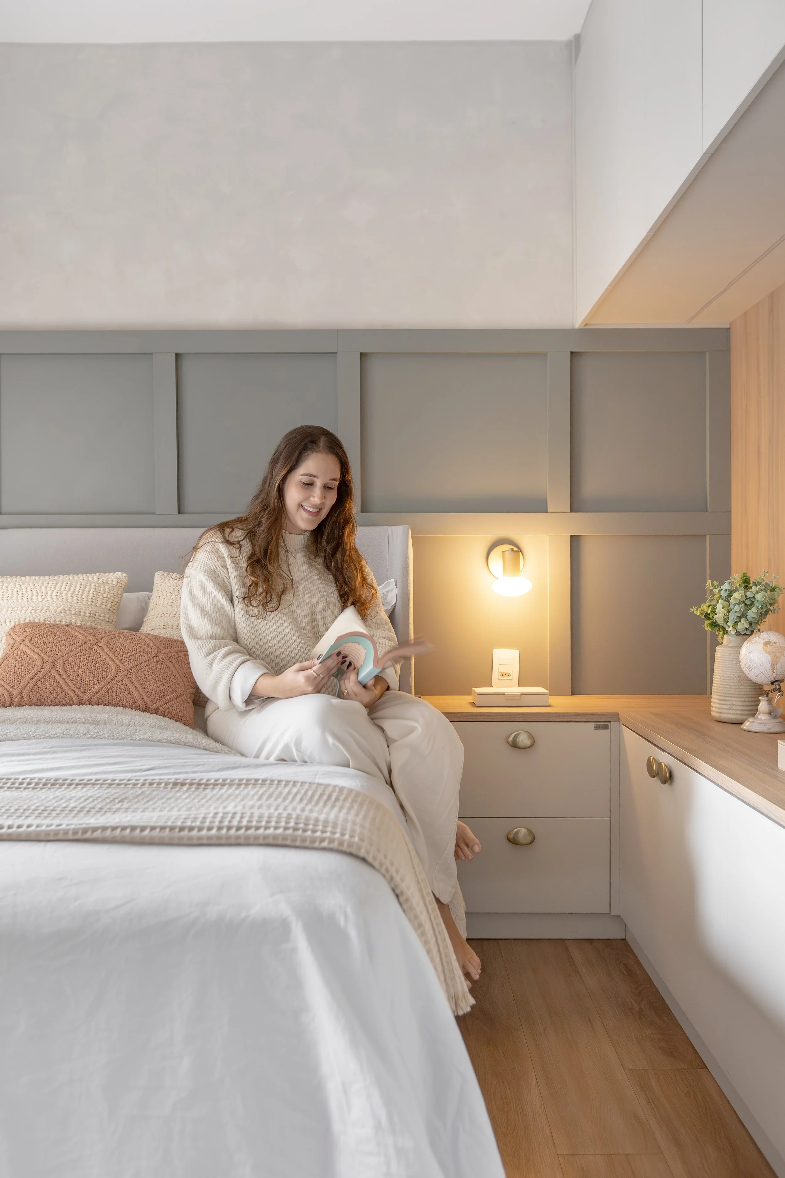 Mulher sorridente sentada na cama, lendo um livro, em um quarto bem iluminado com uma cabeceira de parede cinza e decoração moderna, incluindo uma luminária e uma planta.
