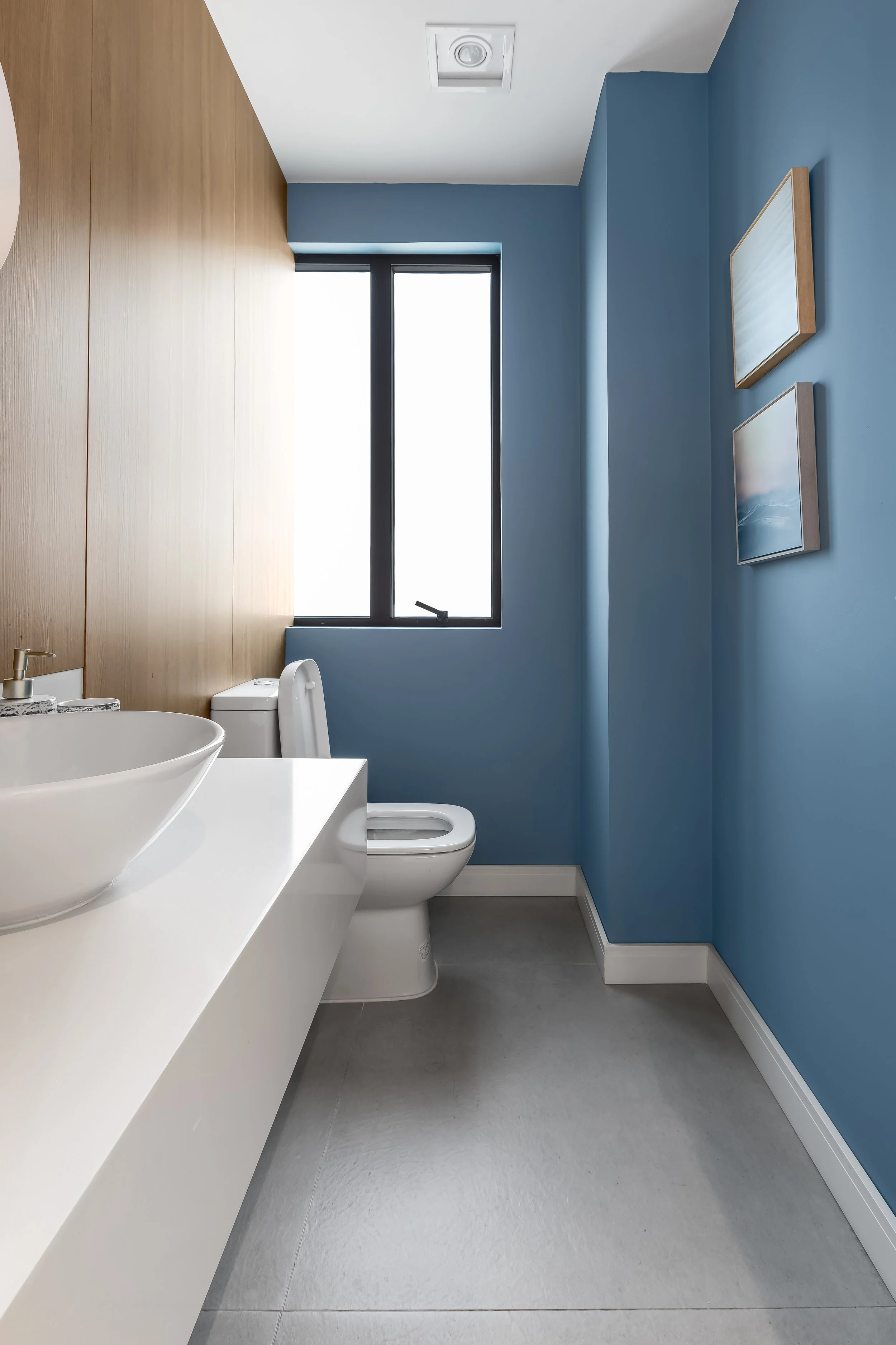 Banheiro com paredes azul claro, janela de vidro, pia de bancada branca com vaso de cerâmica, toalheiro e quadros na parede.