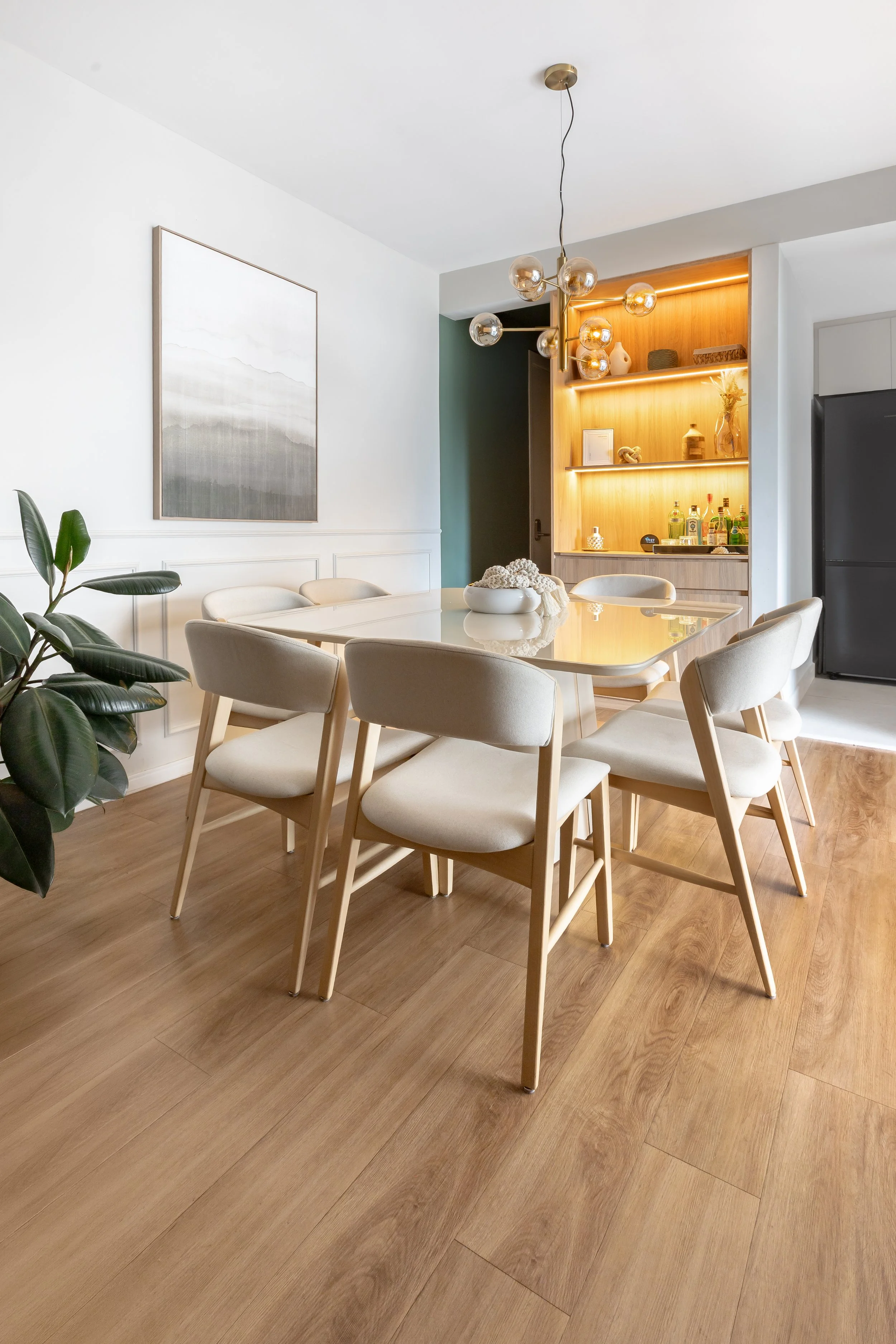 Sala de jantar com mesa branca, oito cadeiras brancas, planta de folhas grandes, parede branca, quadro decorativo minimalista, prateleira de madeira com iluminação, objetos decorativos e bebidas, luminária moderna com globos de vidro.