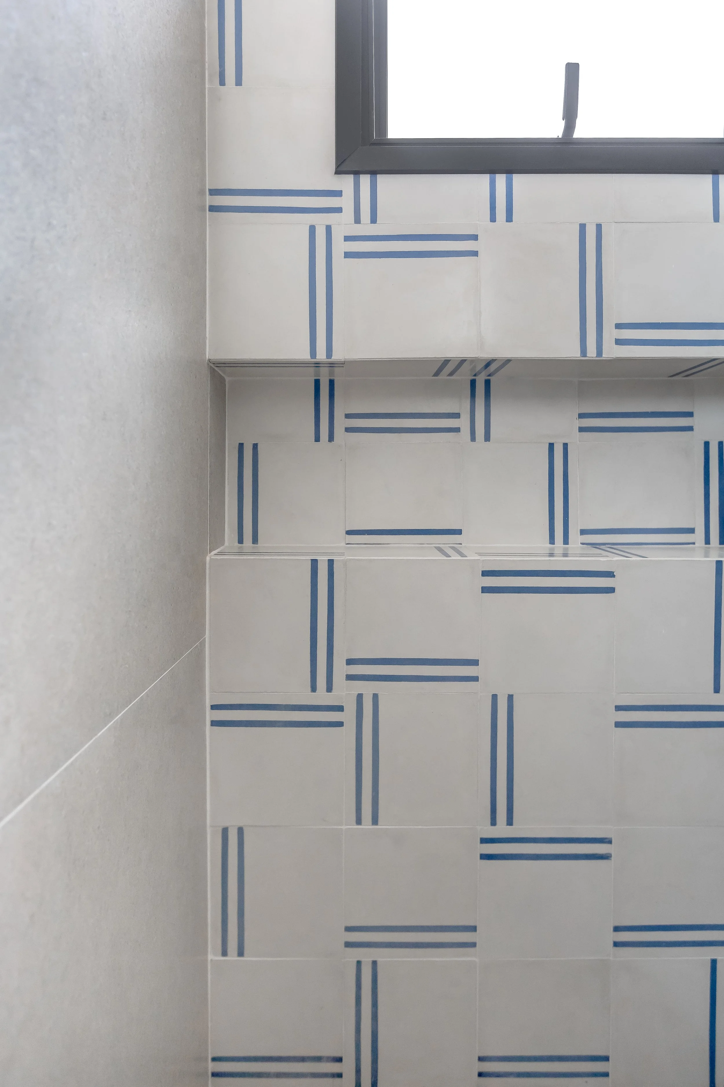 Parede de azulejos branco com detalhes em linhas azuis, perto de uma janela de vidro com moldura cinza.