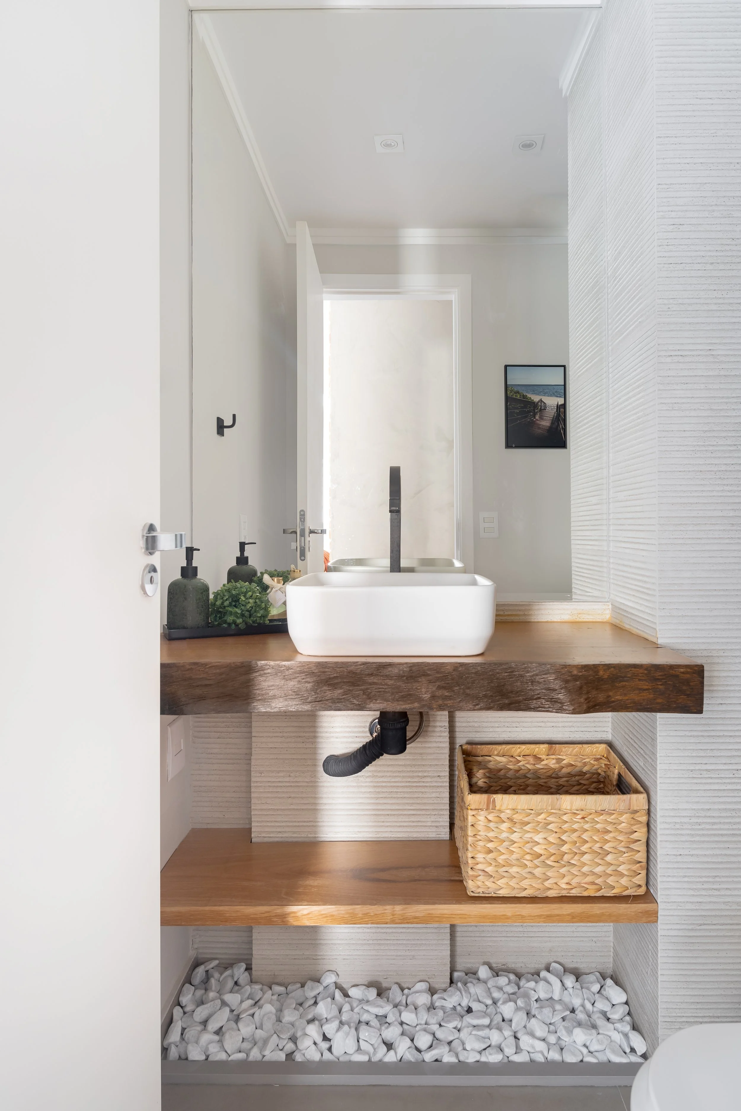 Banheiro com pia de tampo de madeira, cuba branca, vaso sanitário e decoração com plantas e pedra branca no chão.