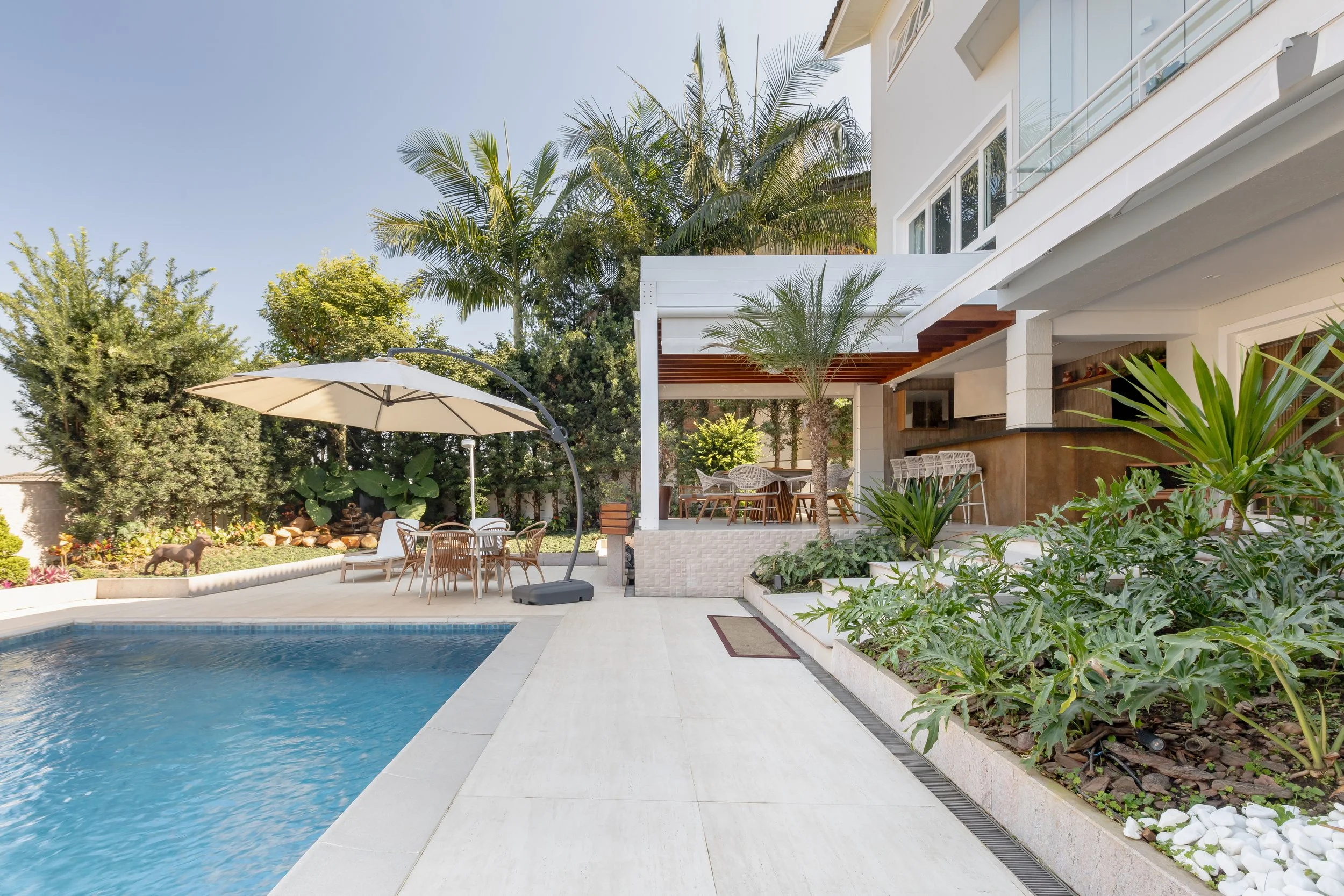 Área de piscina com espreguiçadeiras, guarda-sol, jardim com árvores e plantas, varanda de uma casa moderna com espaço de churrasqueira.