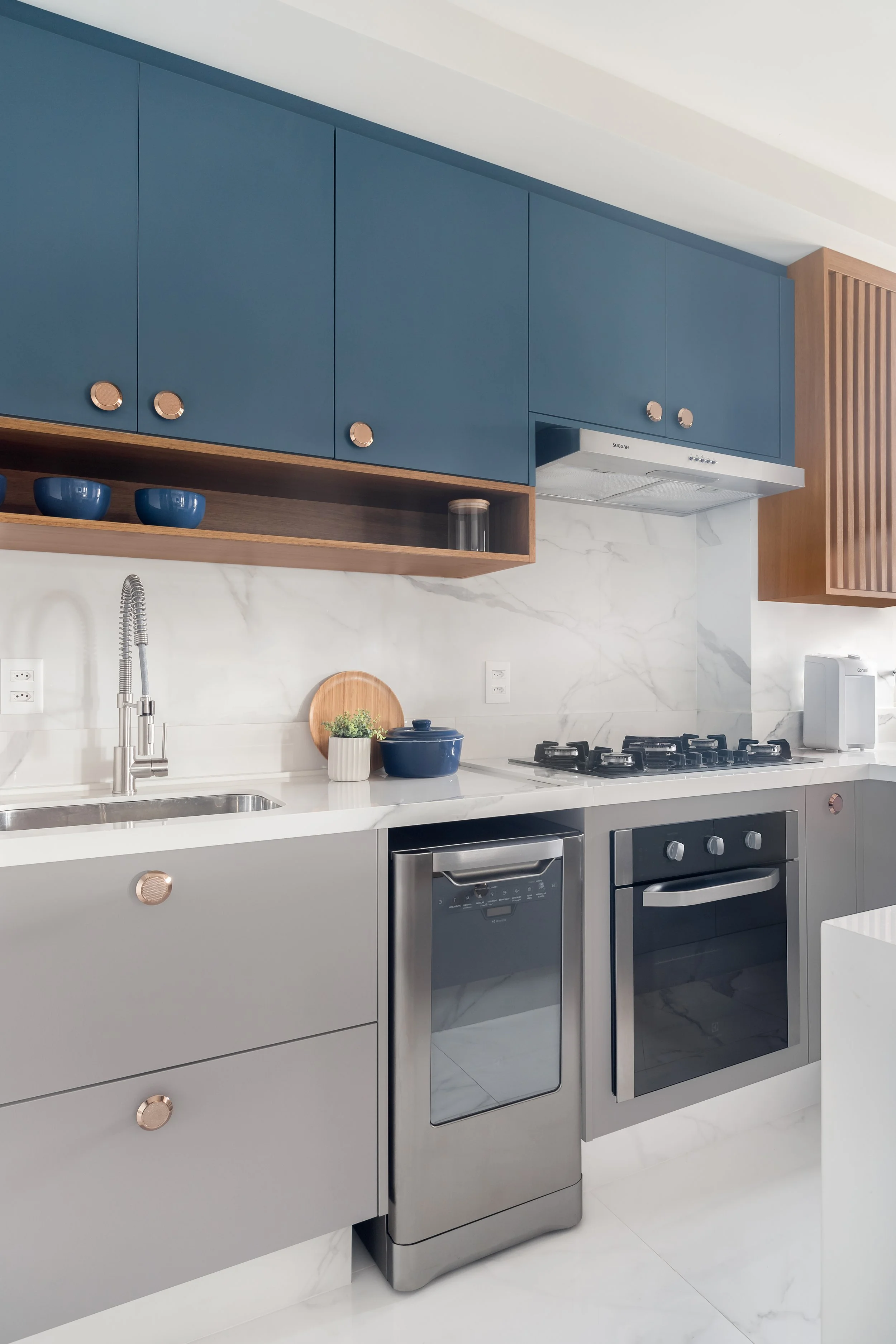 Cozinha moderna com armários azuis, bancada de mármore, eletrodomésticos de aço inox, incluindo fogão, forno e lava-louças, com detalhes em madeira e decoração simples.