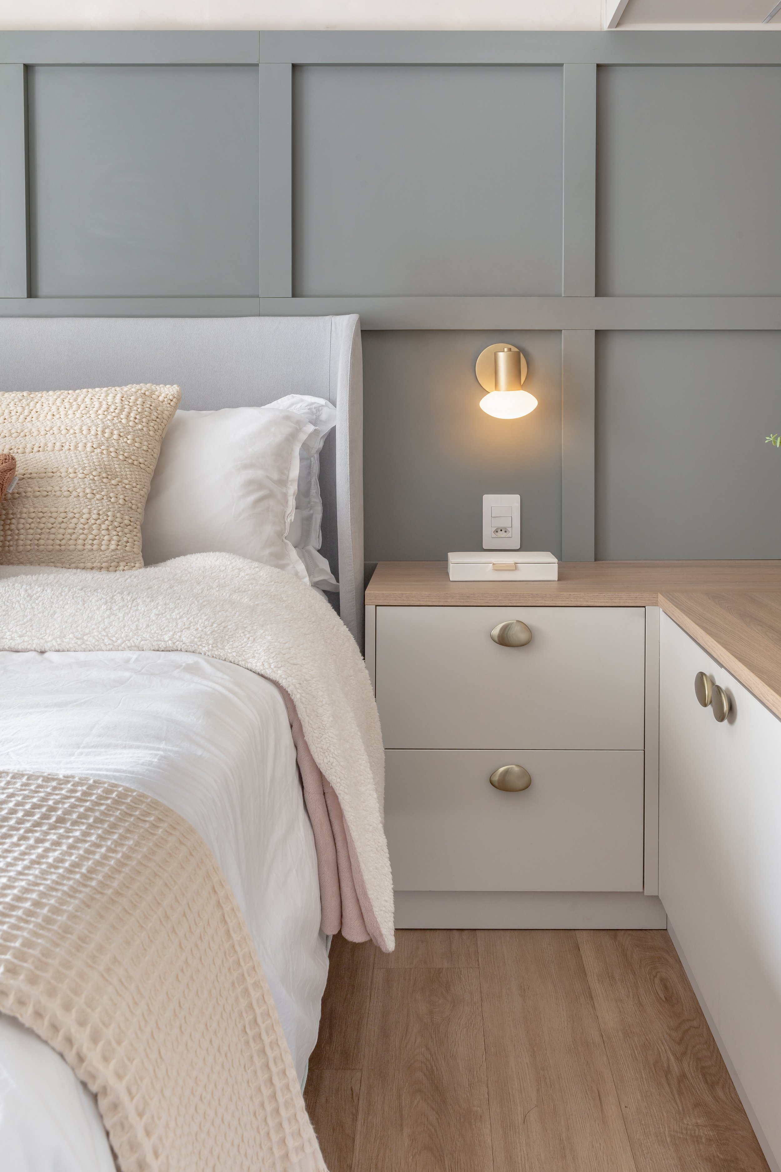 Cama de casal com travesseiro bege, manta bege e roupa de cama branca em um quarto com parede cinza e cabeceira estofada. Ao lado da cama, cômoda branca com gavetas e detalhes em dourado, com luminária dourada acesa.