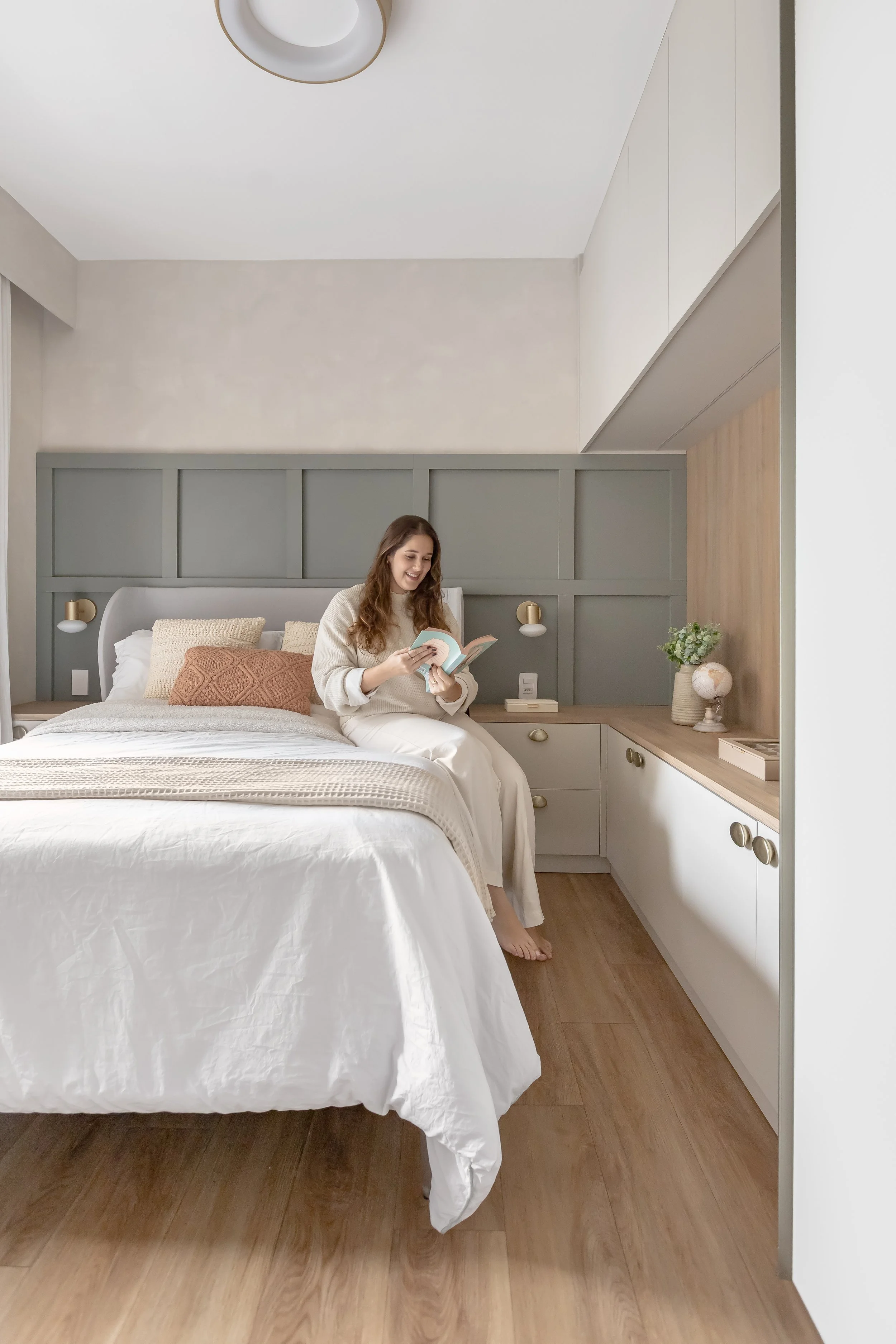 Mulher de cabelo castanho claro sentado na cama lendo um livro, com roupa de cor bege ou branca, em um quarto bem decorado com armários brancos, parede cinza, luminárias de parede douradas, uma pequena planta e objetos decorativos ao lado da cama.