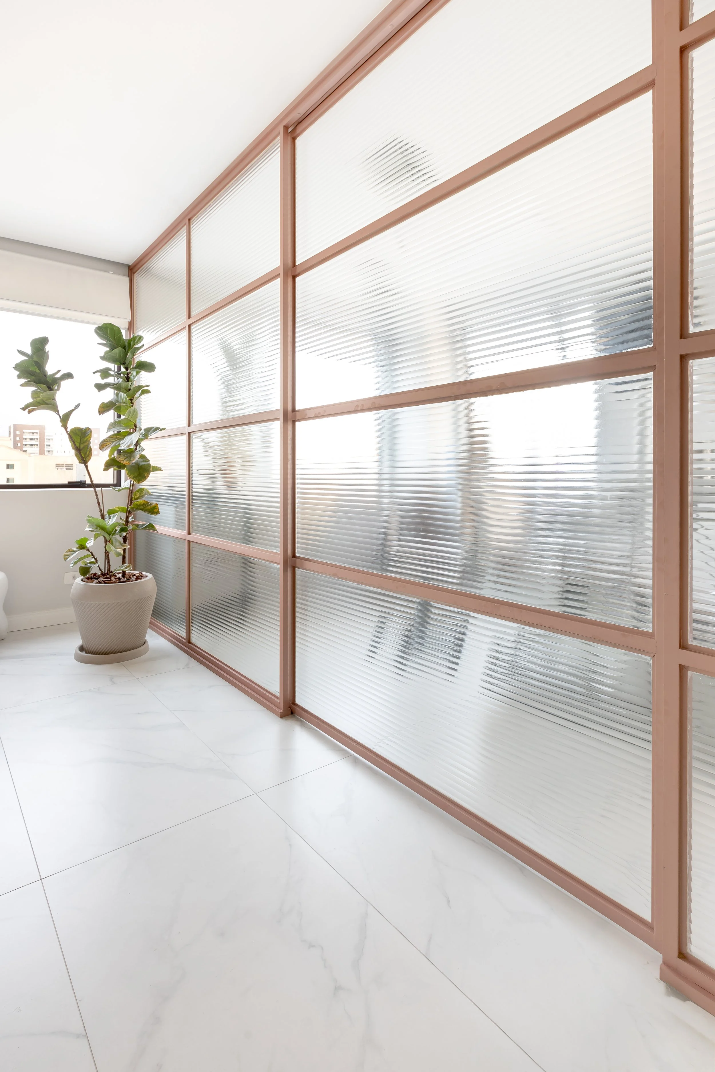 Divisão de ambiente com parede de vidro fosco com moldura rosa, em um espaço com piso de mármore branco e planta em vaso decorativo.