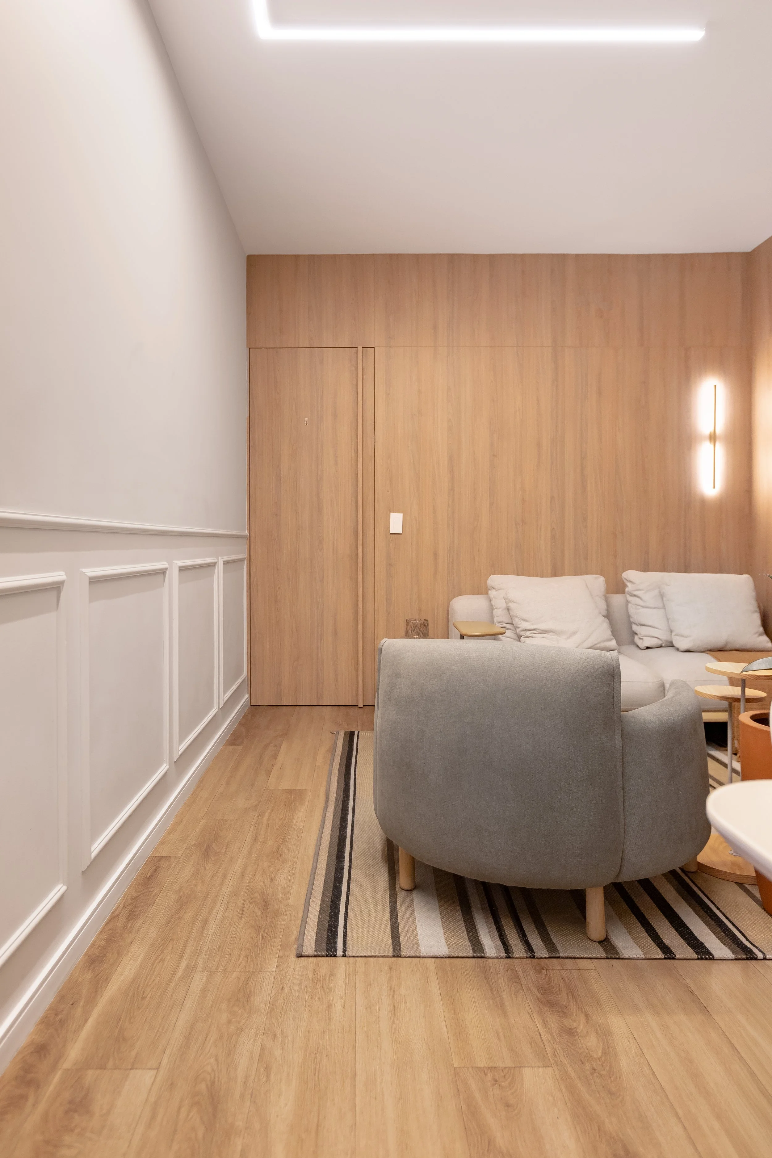 Sala de estar com piso de madeira, parede de madeira clara e paredes brancas, sofá branco, duas poltronas cinza, mesa de canto de madeira e um tapete listrado em tons de bege, preto e branco, com iluminação moderna na parede.
