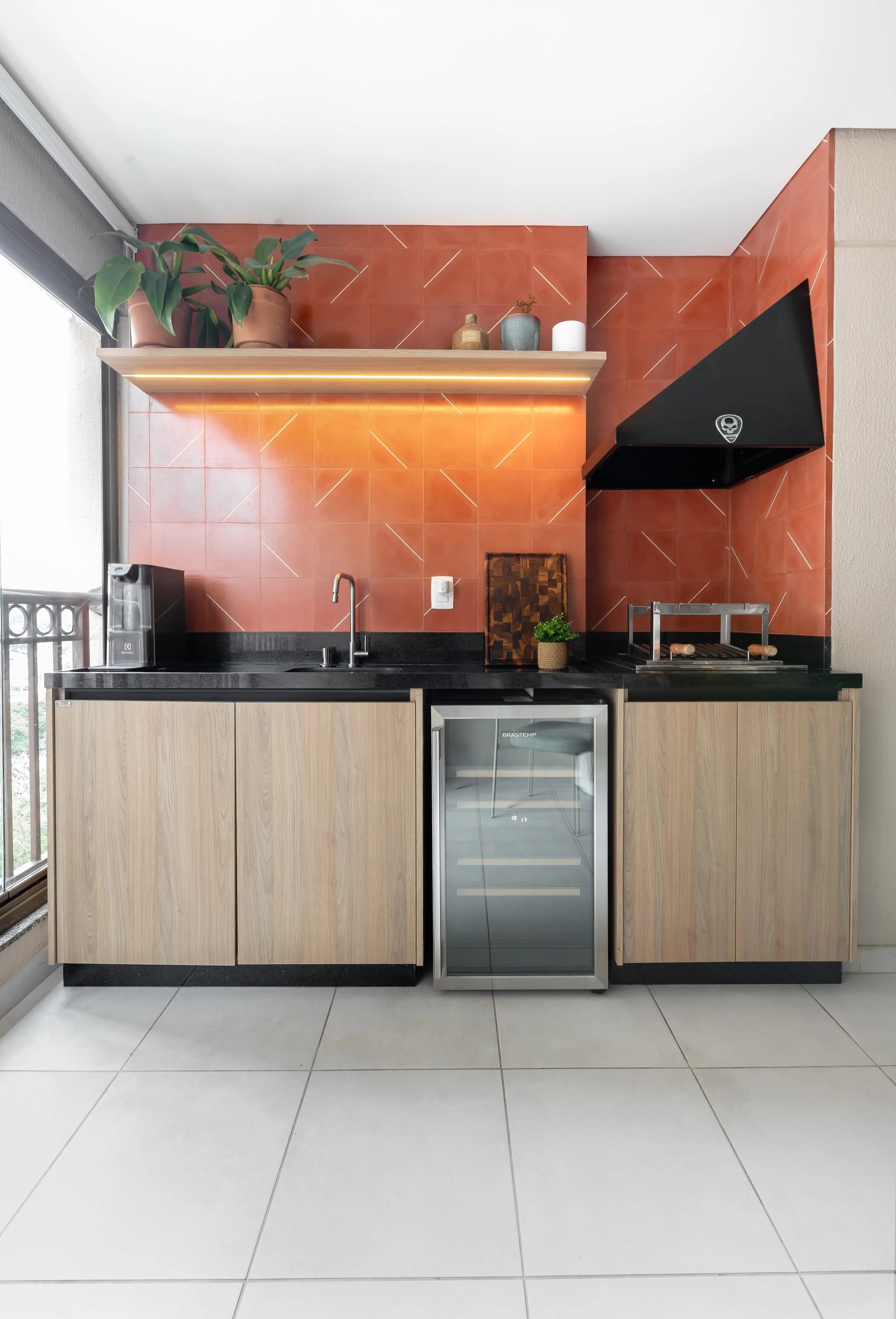 Cozinha moderna com armários de madeira, balcão preto, micro-ondas, adega, pia com torneira, parede de azulejos vermelhos, prateleira com plantas, utensílios de cozinha e janela ao lado.