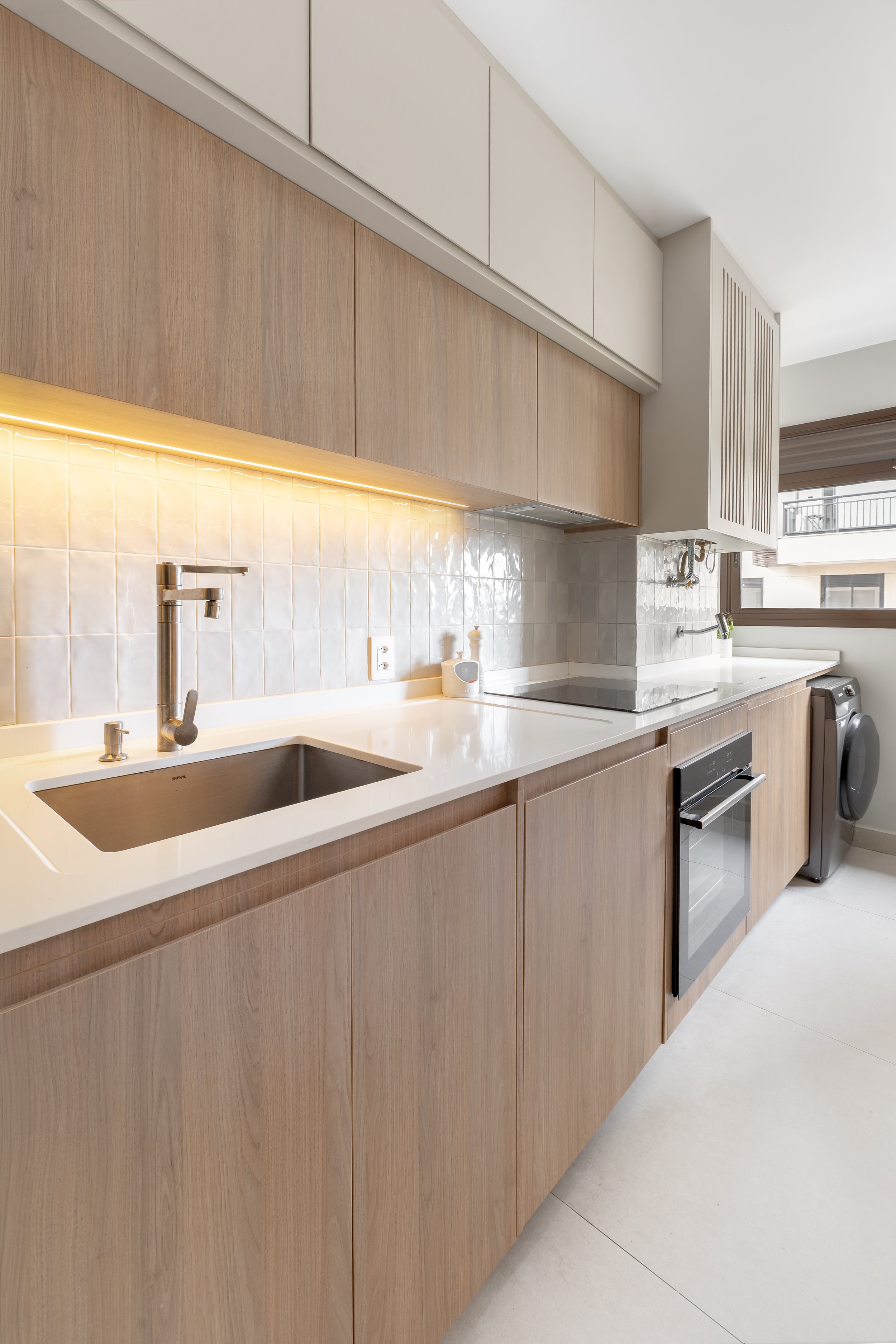 Cozinha moderna com armários de madeira clara, pia de aço inox, backsplash de azulejos brancos e eletrodomésticos de inox, ao lado de uma janela com persiana e varandão.