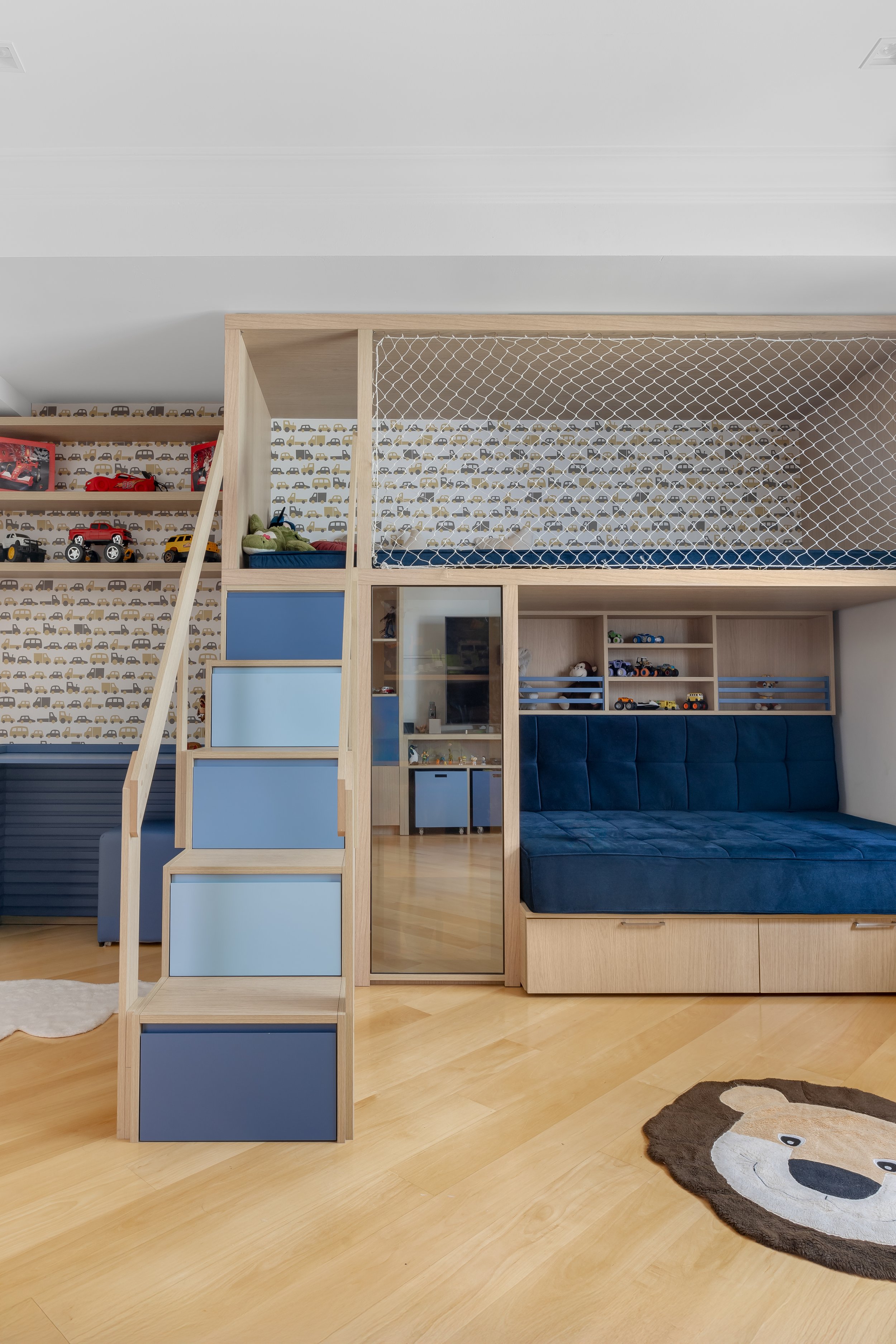 Quarto infantil com cama no mezanino, escada com gavetas coloridas, parede decorada com papel de parede de carros, tapete com rosto de urso, e espaço de brincar com brinquedos.