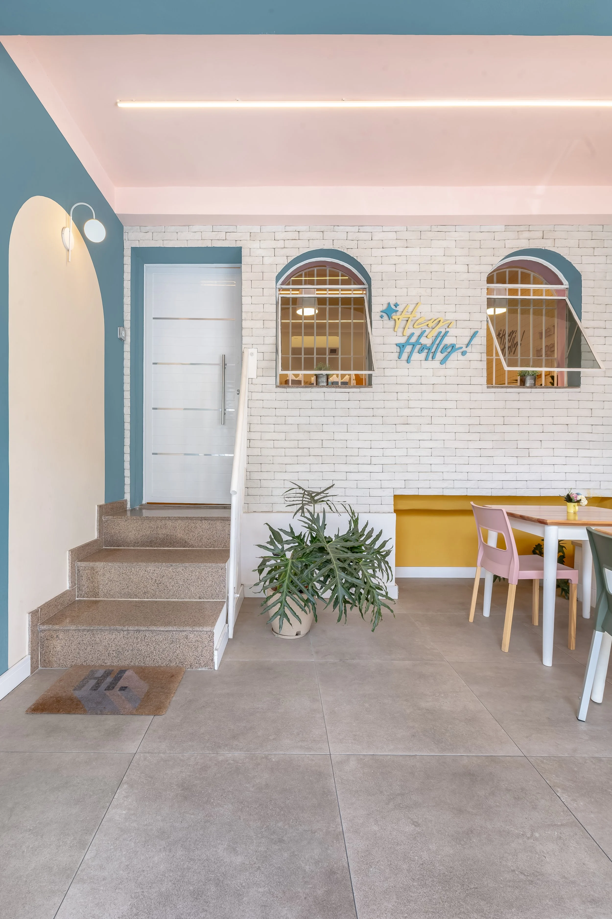 Interior de um espaço decorado com parede de tijolos brancos, uma mesa com cadeiras coloridas, uma planta grande, uma porta branca, uma parede azul e uma placa que diz 'Hey Holly!'.
