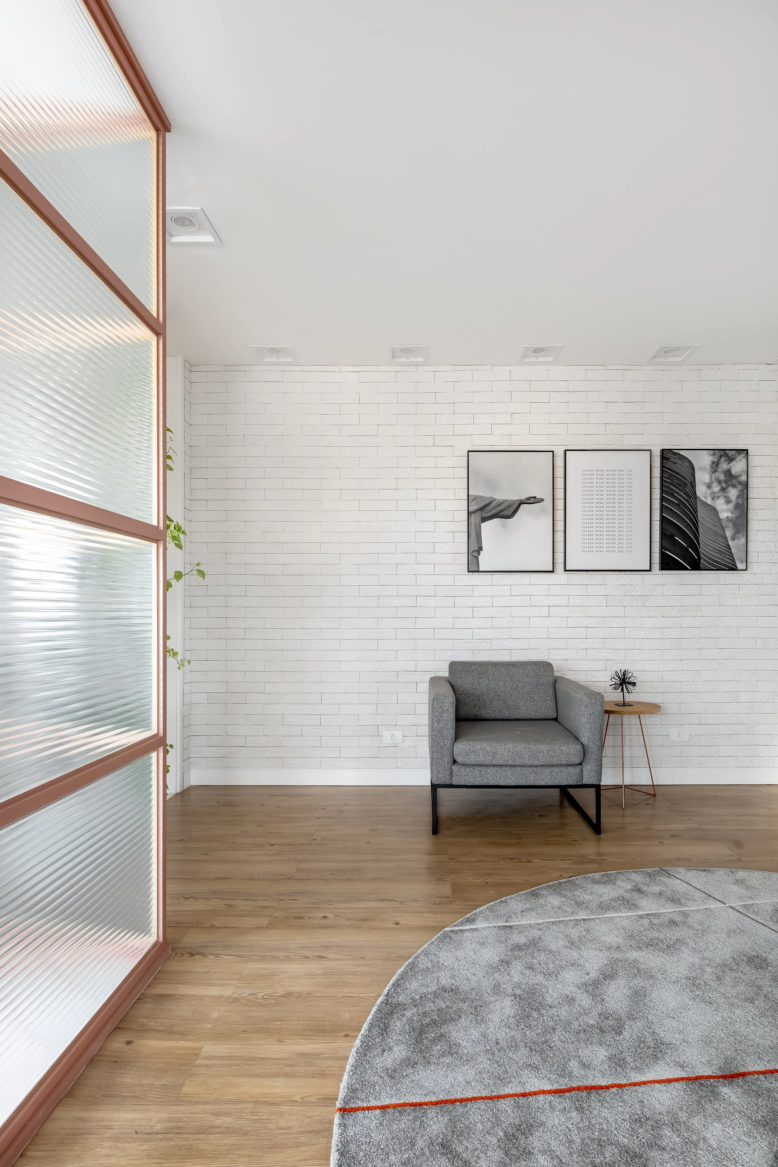 Sala de estar com parede de tijolos brancos, uma poltrona cinza, um quadro com três imagens em preto e branco, uma mesa pequena ao lado da poltrona e uma área de piso de madeira e uma área de carpete cinza redondo.