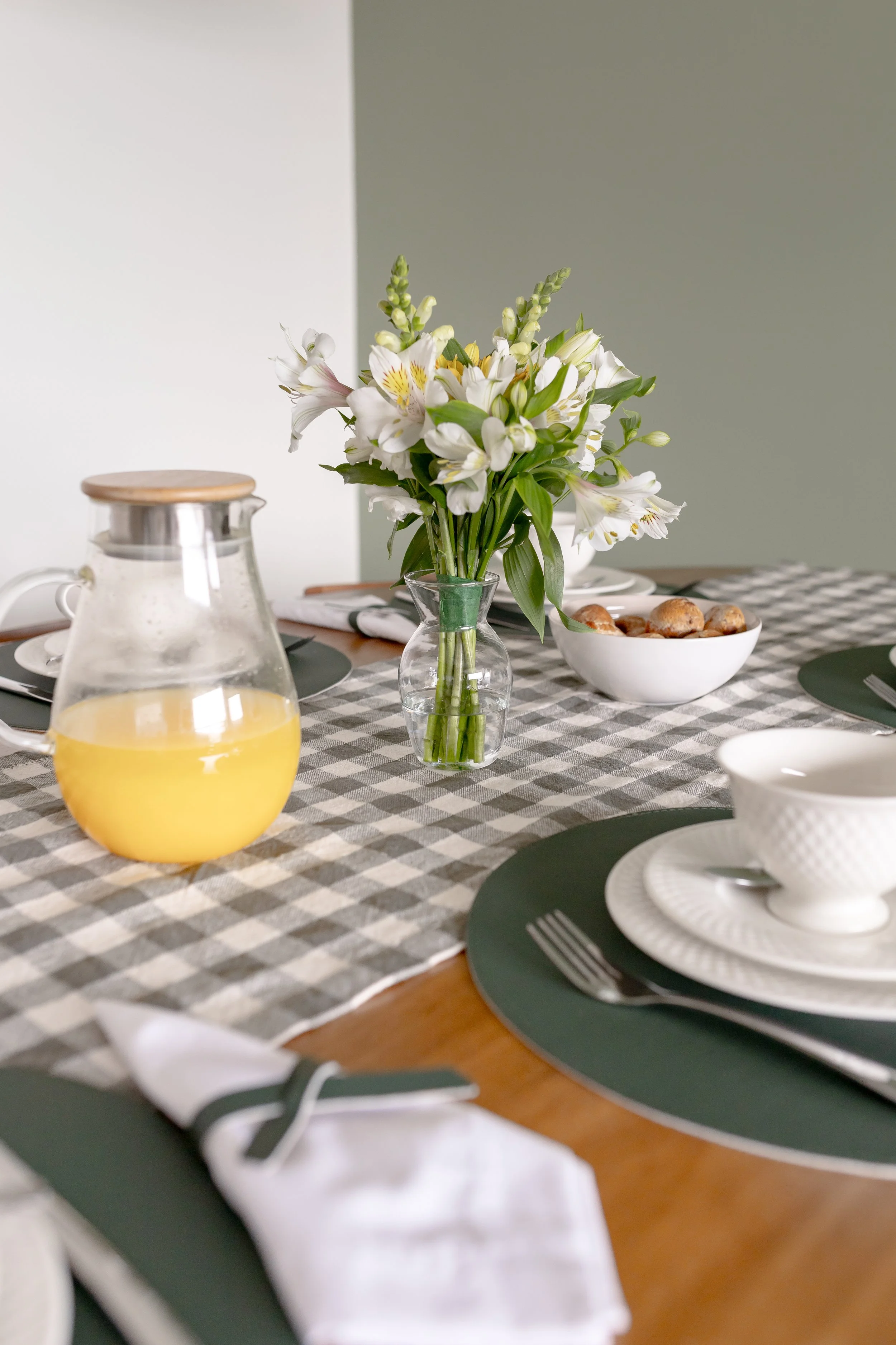 Mesa de jantar decorada com flores brancas em vaso, tigela com pães, panqueca de suco de laranja, copos e pratos brancos sobre passadeiras verdes em uma mesa de madeira.
