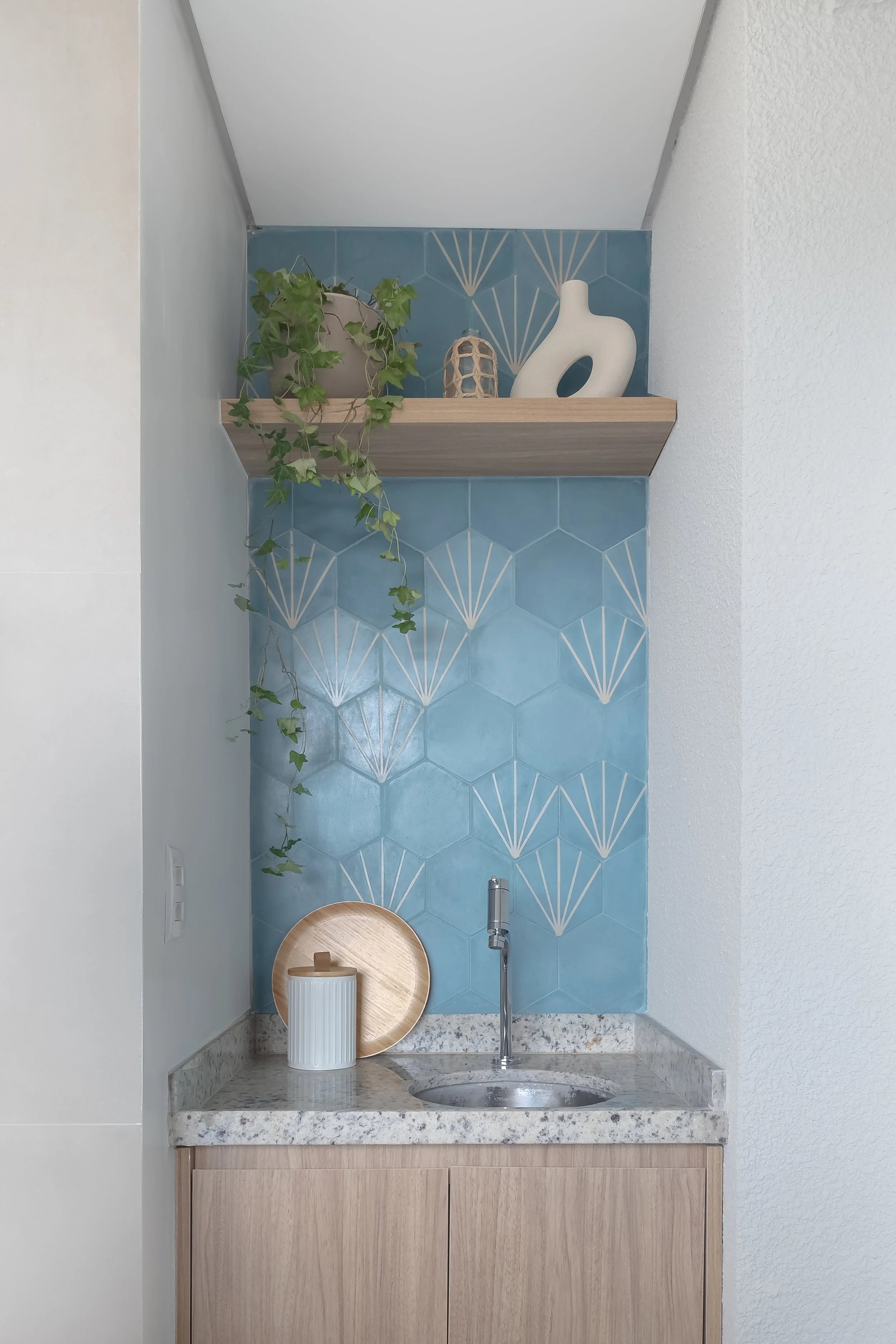 Pequena pia com bancada de granito, armário de madeira, parede com azulejos azuis hexagonais com detalhes brancos, vaso com plantas e objetos decorativos na prateleira.