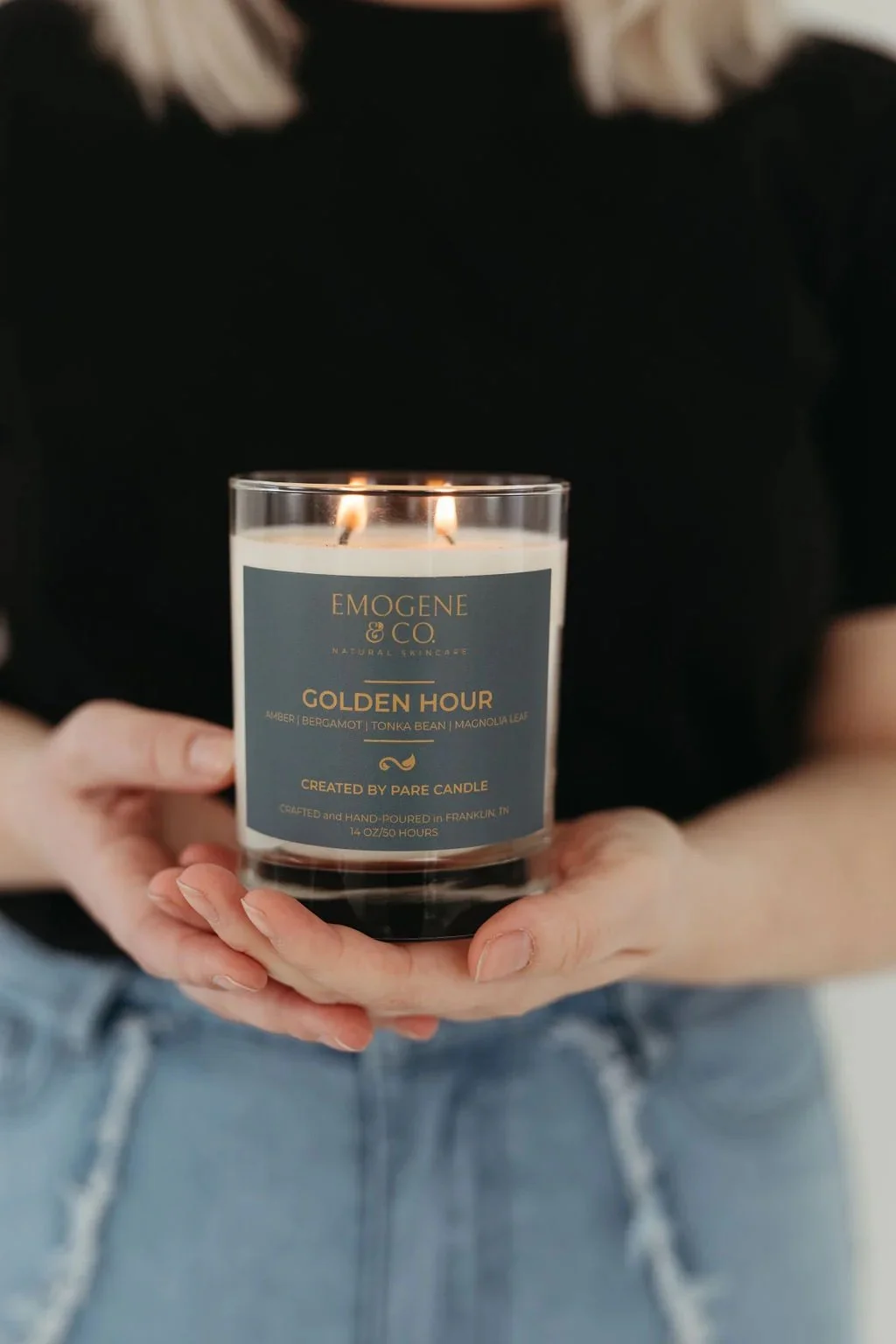 Person holding a lit scented candle labeled 'Golden Hour' by Emogene & Co, featuring notes of amber, bergamot, tonka bean, and magnolia leaf.