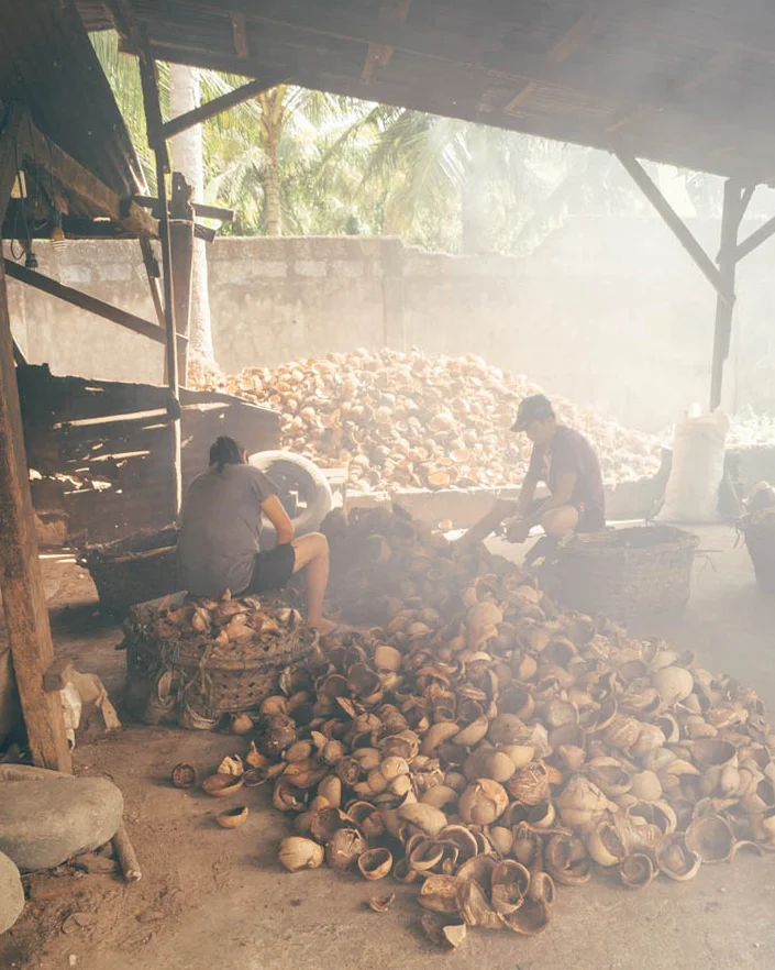 two workers sat in a dark hazy roofed outdoor workspace amongst dozens of removed coconut shells on the floor