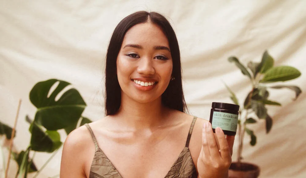 girl standing in front of beige backdrop and plants holding CBD coconut oil