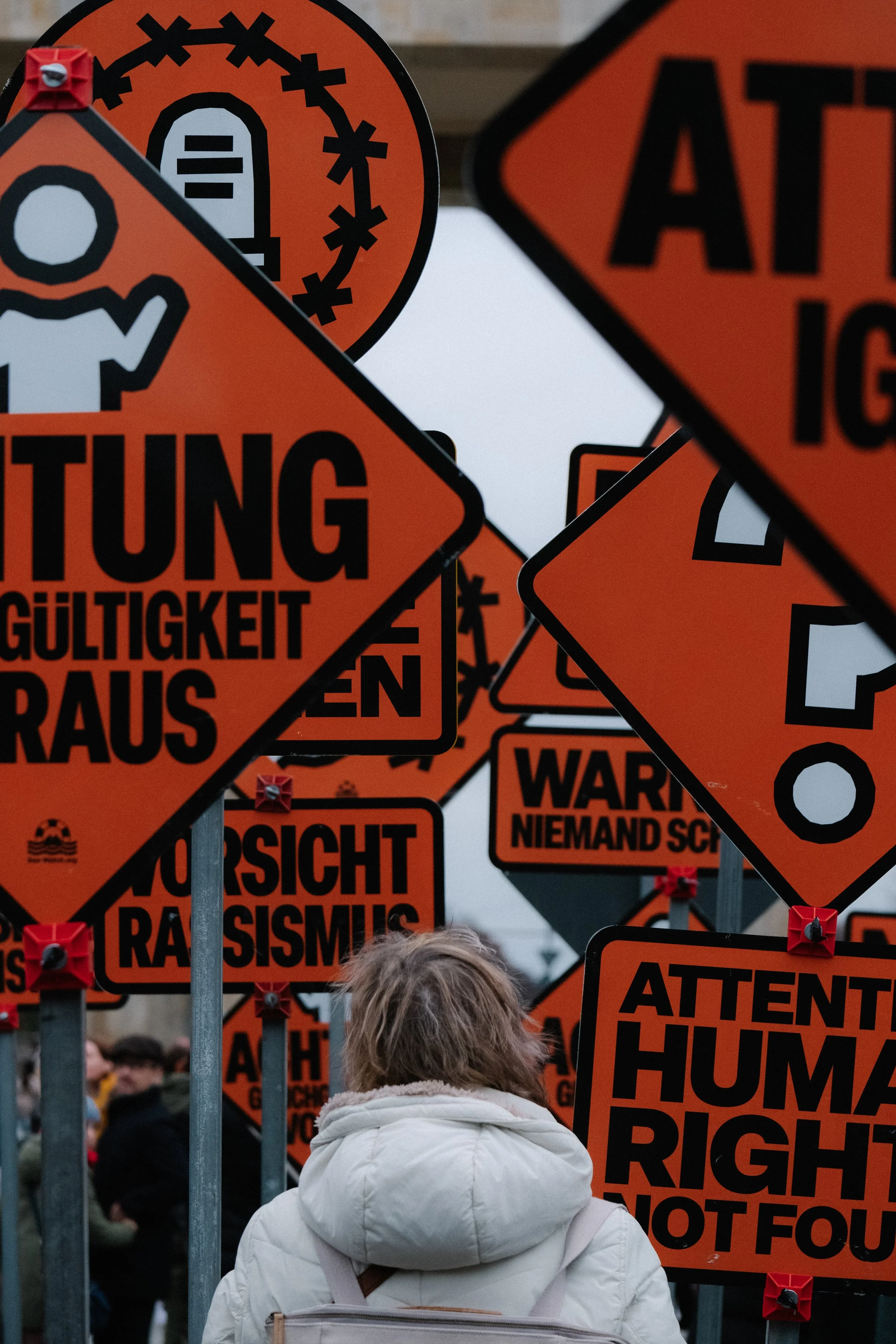 A person wearing a white coat standing among numerous protest signs with bold black text on orange backgrounds, some with symbols, containing messages in English and German, protesting human rights and warning about racism.