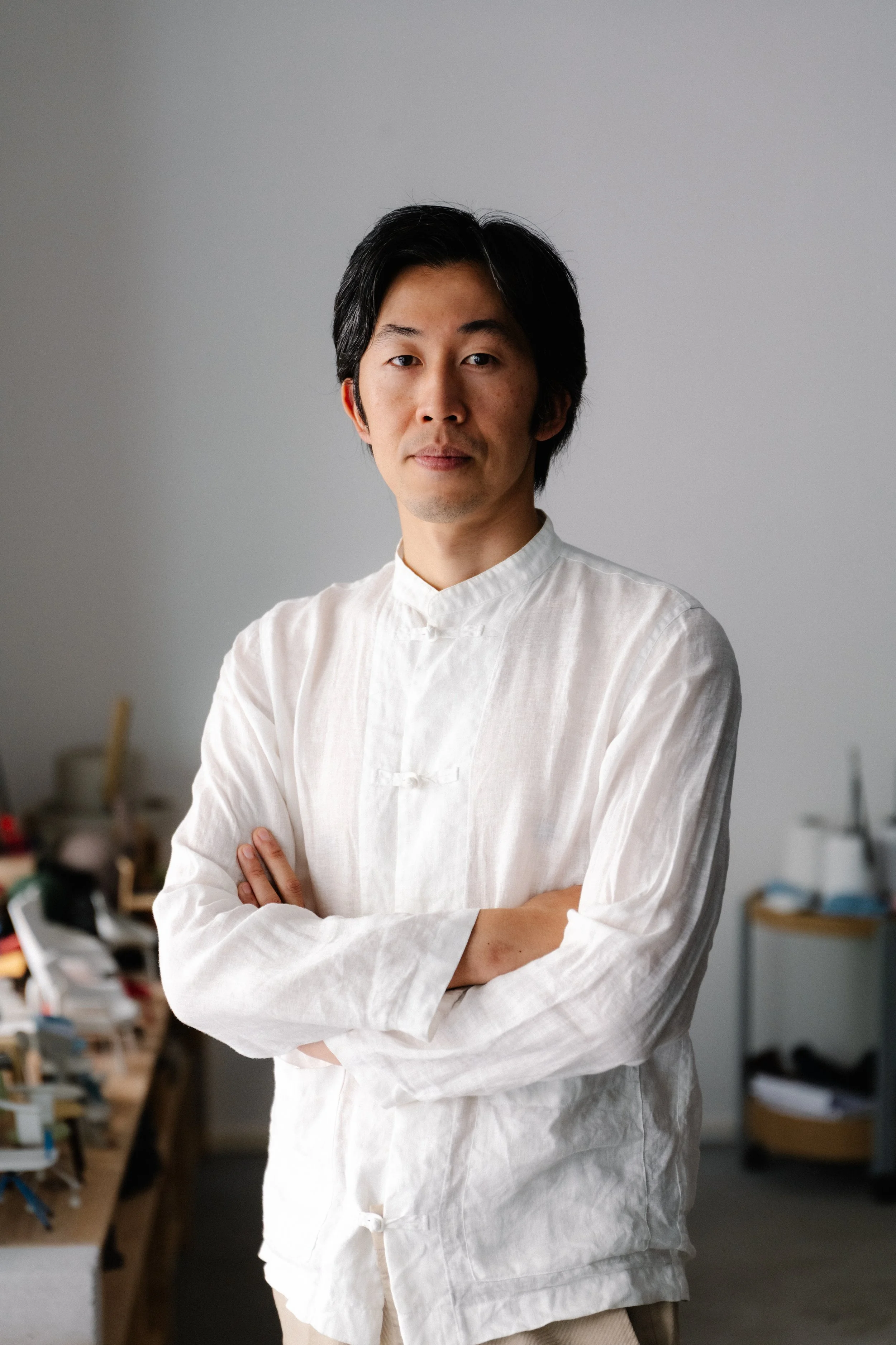 Man with black hair and light skin, wearing a white traditional Chinese shirt, standing with arms crossed in an indoor space with blurred objects in the background.