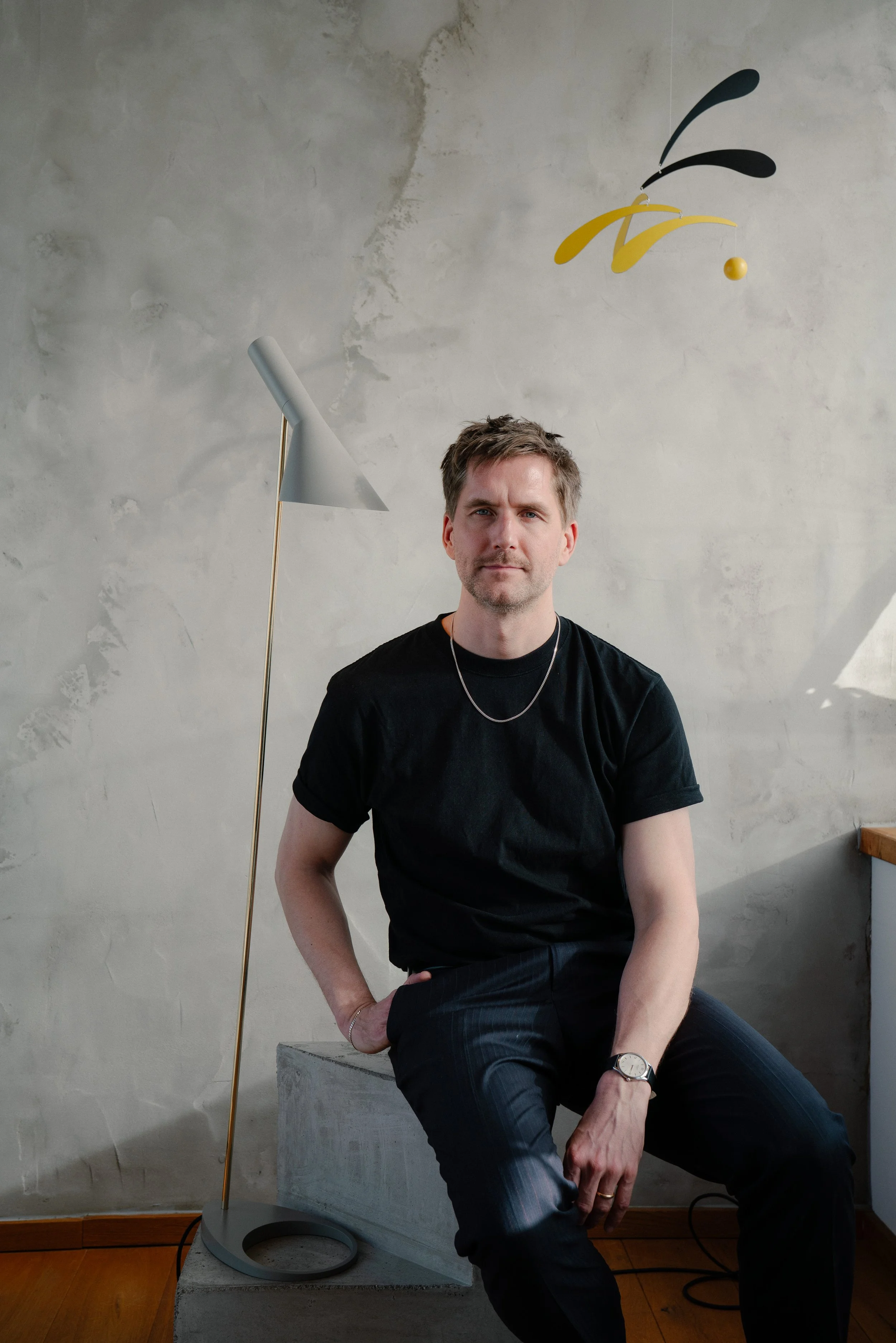 A man with short brown hair and a beard sitting on a concrete block in a room with a textured gray wall. He's wearing a black t-shirt, dark striped pants, a watch on his left wrist, a necklace, and a ring on his right hand. Behind him, there is a modern floor lamp with a light gray shade and a yellow and black mobile hanging from the ceiling.