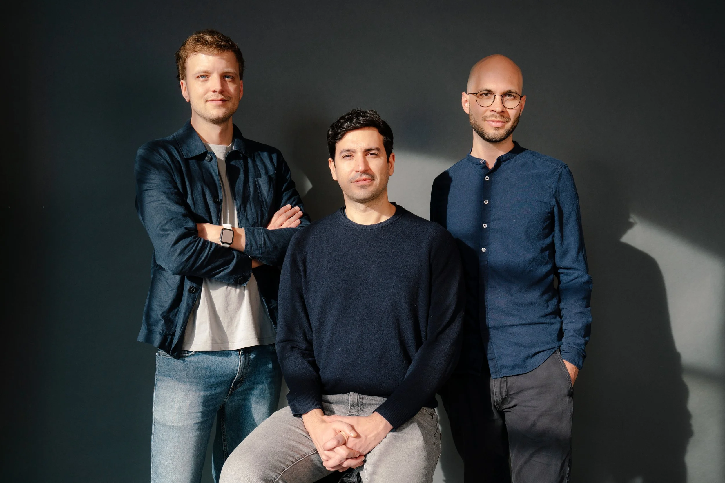 Three men standing against a dark gray wall, posing for a photo. The man on the left has short brown hair, wearing a dark blue jacket, white t-shirt, and jeans. The man in the middle has black hair, wearing a dark sweater and gray pants, sitting on a stool. The man on the right has a shaved head, wearing glasses, a blue shirt, and dark pants.