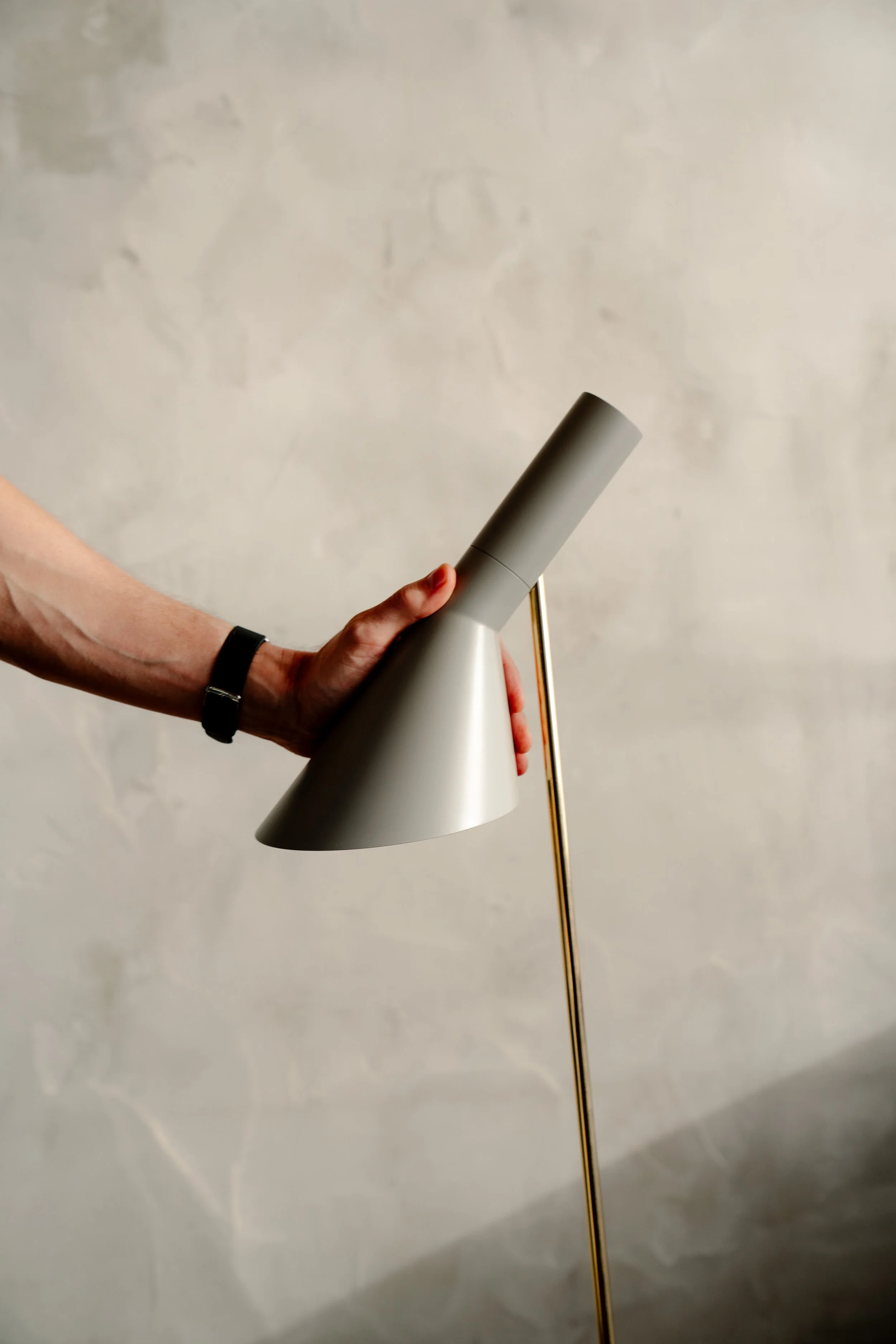 A person holding a modern, cone-shaped, gray desk lamp with a gold stand against a plain, textured beige wall.