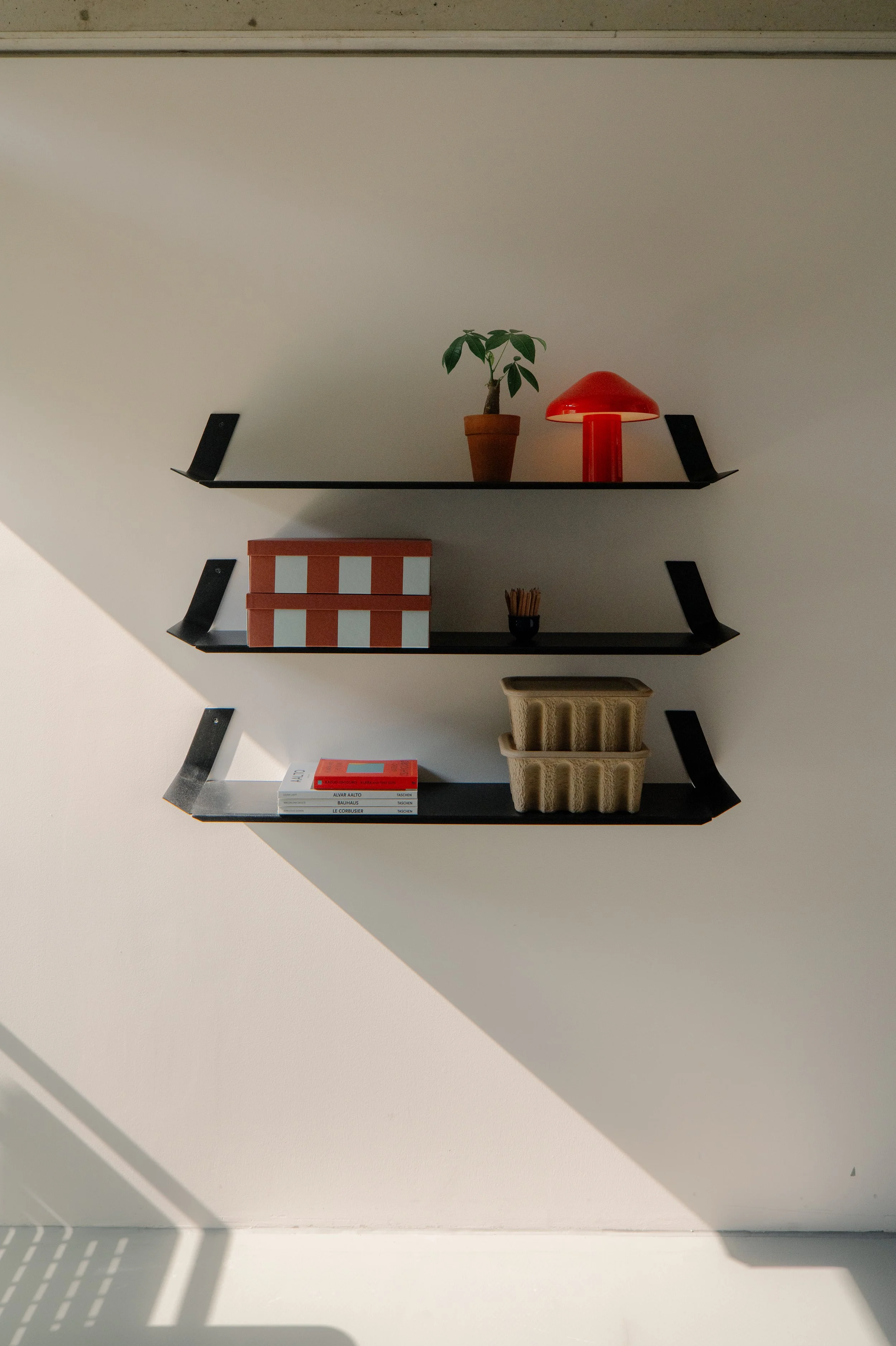 Three black wall shelves with decorative items like a small potted plant, red lamp, boxes, sticks, books, and plant baskets, mounted on a white wall with sunlight casting shadows.