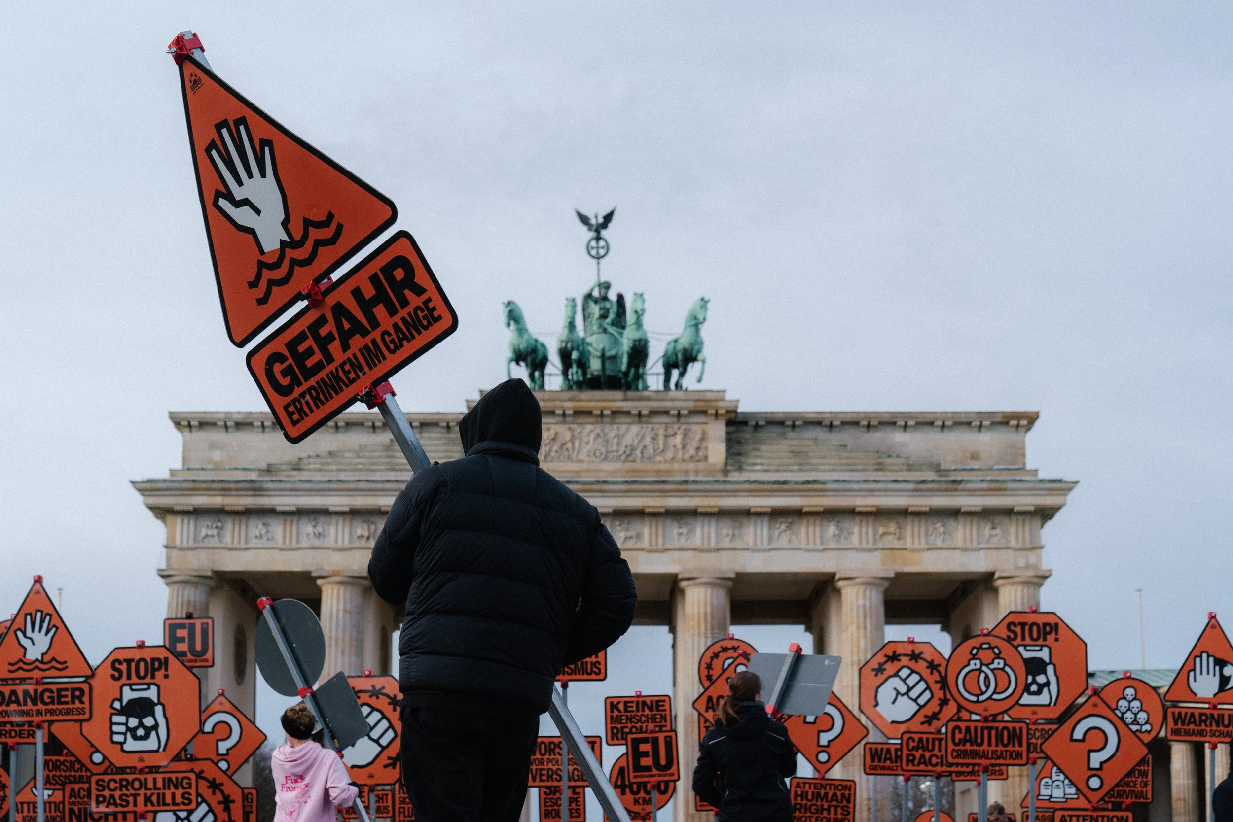 Protestors holding orange warning signs related to environmental and social issues in front of the Brandenburg Gate in Berlin, Germany.