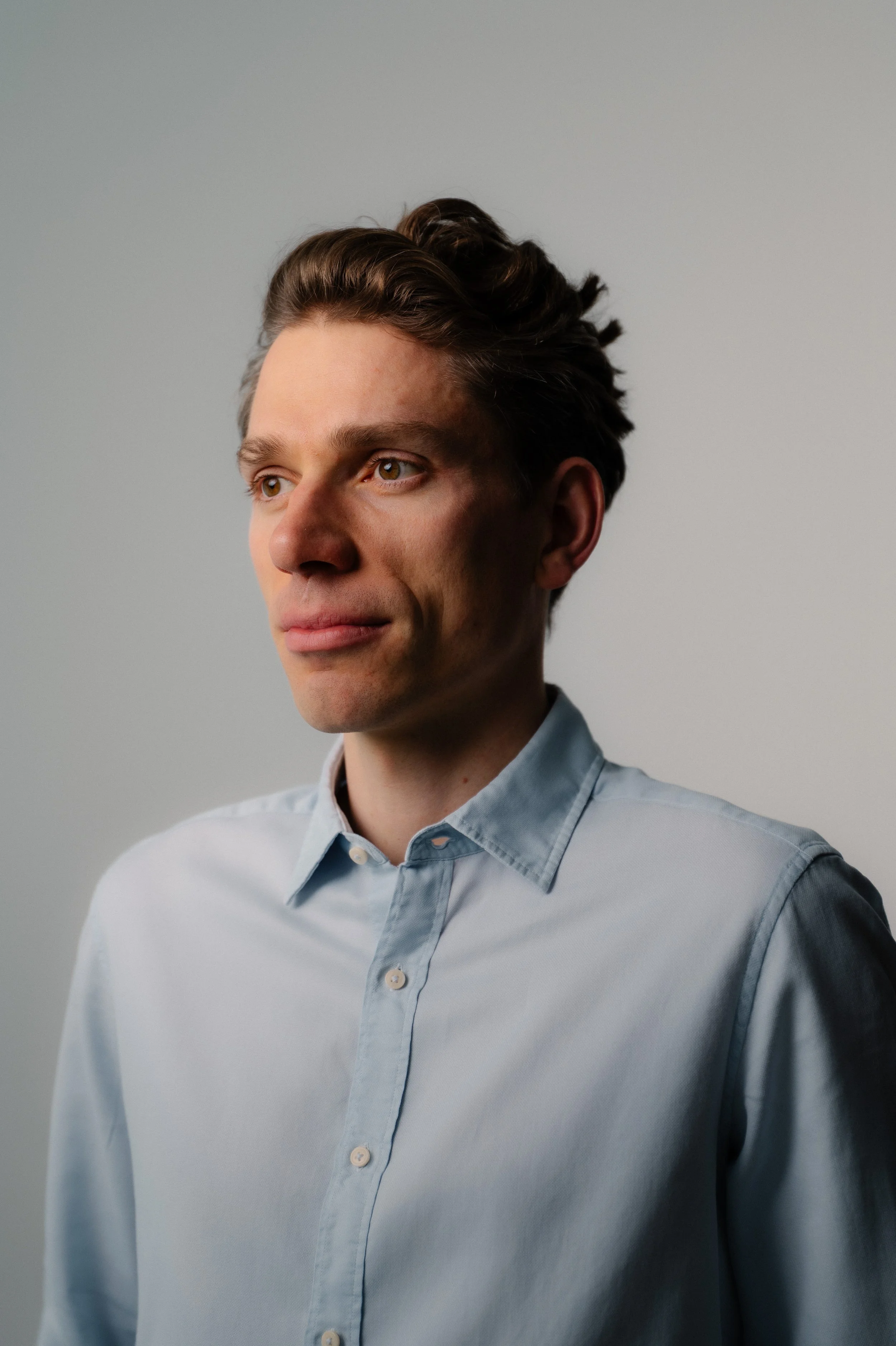 A young man with light skin and styled brown hair, wearing a light blue button-up shirt, looking thoughtfully to the side against a plain gray background.