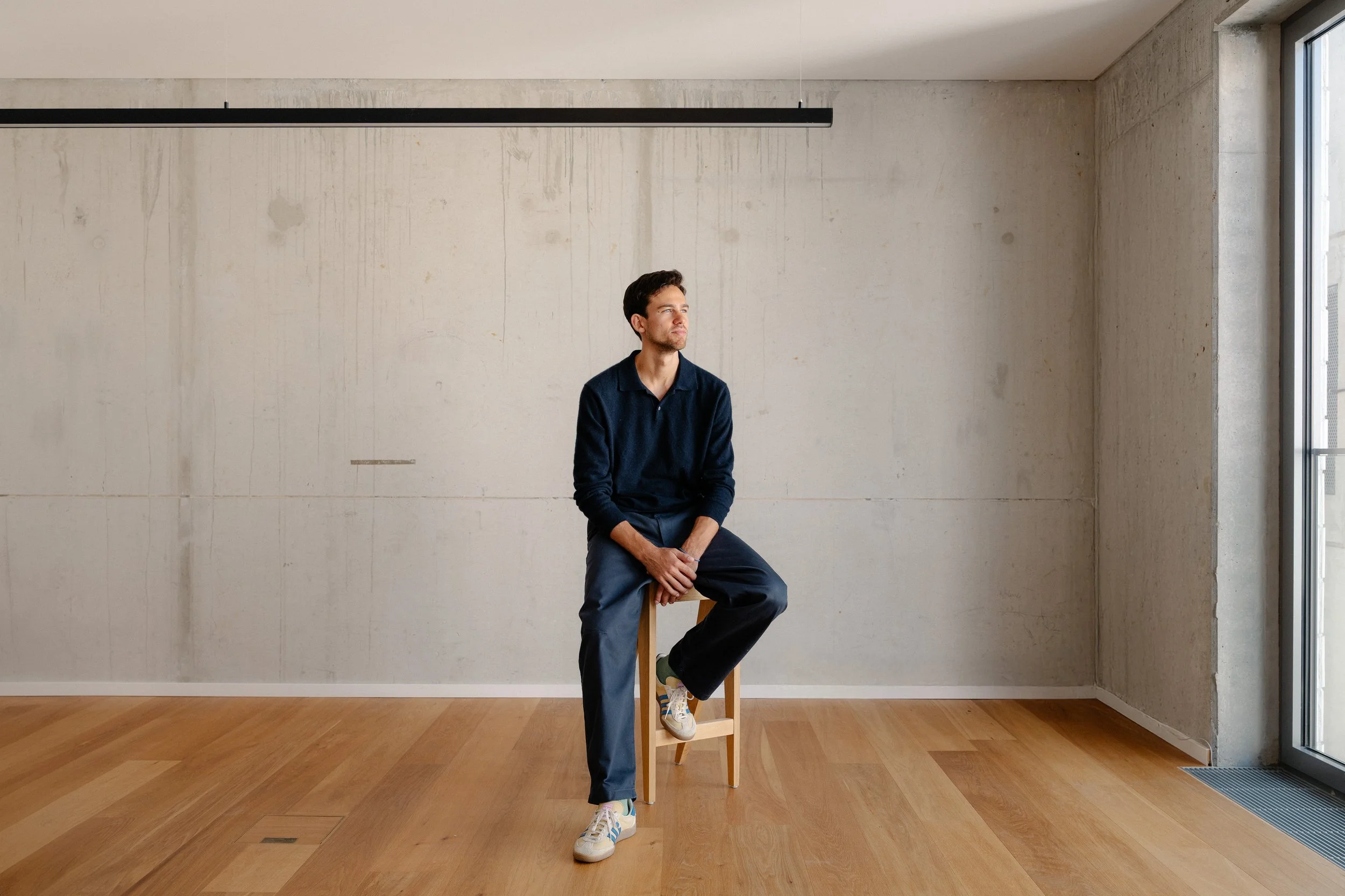 A man sitting on a wooden stool in a minimalist room with a concrete wall and wooden floor, near a large window.