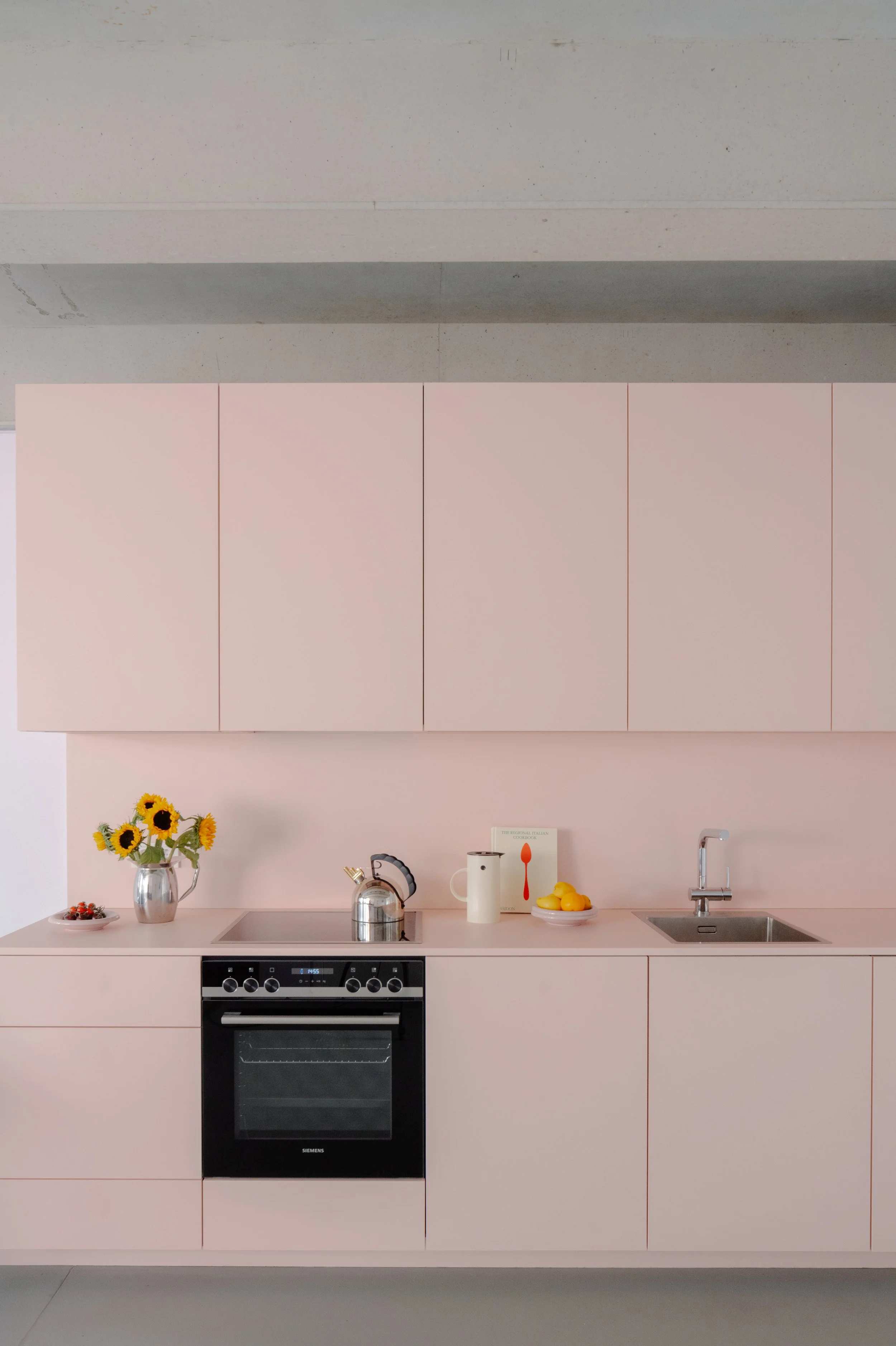 Modern kitchen with light pink cabinets, a stainless steel oven, a kettle, a vase with sunflowers, a bowl of lemons, and a sink on the right.