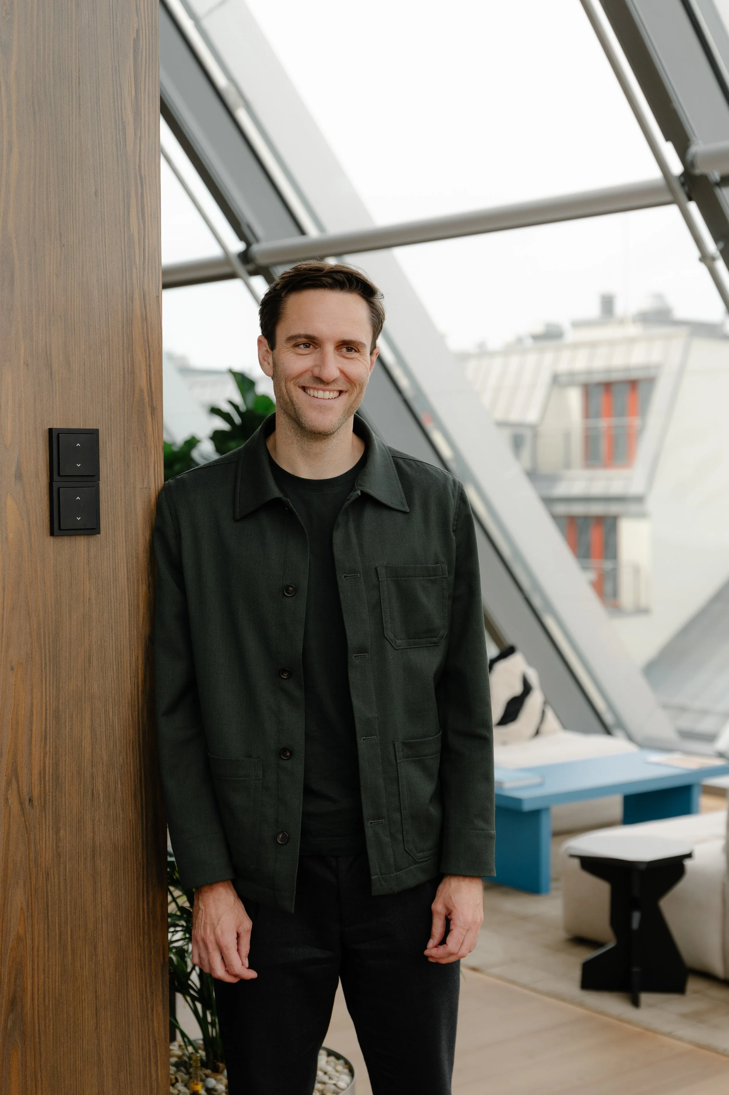 A smiling man standing indoors near a wooden wall with a modern building with large glass windows in the background.