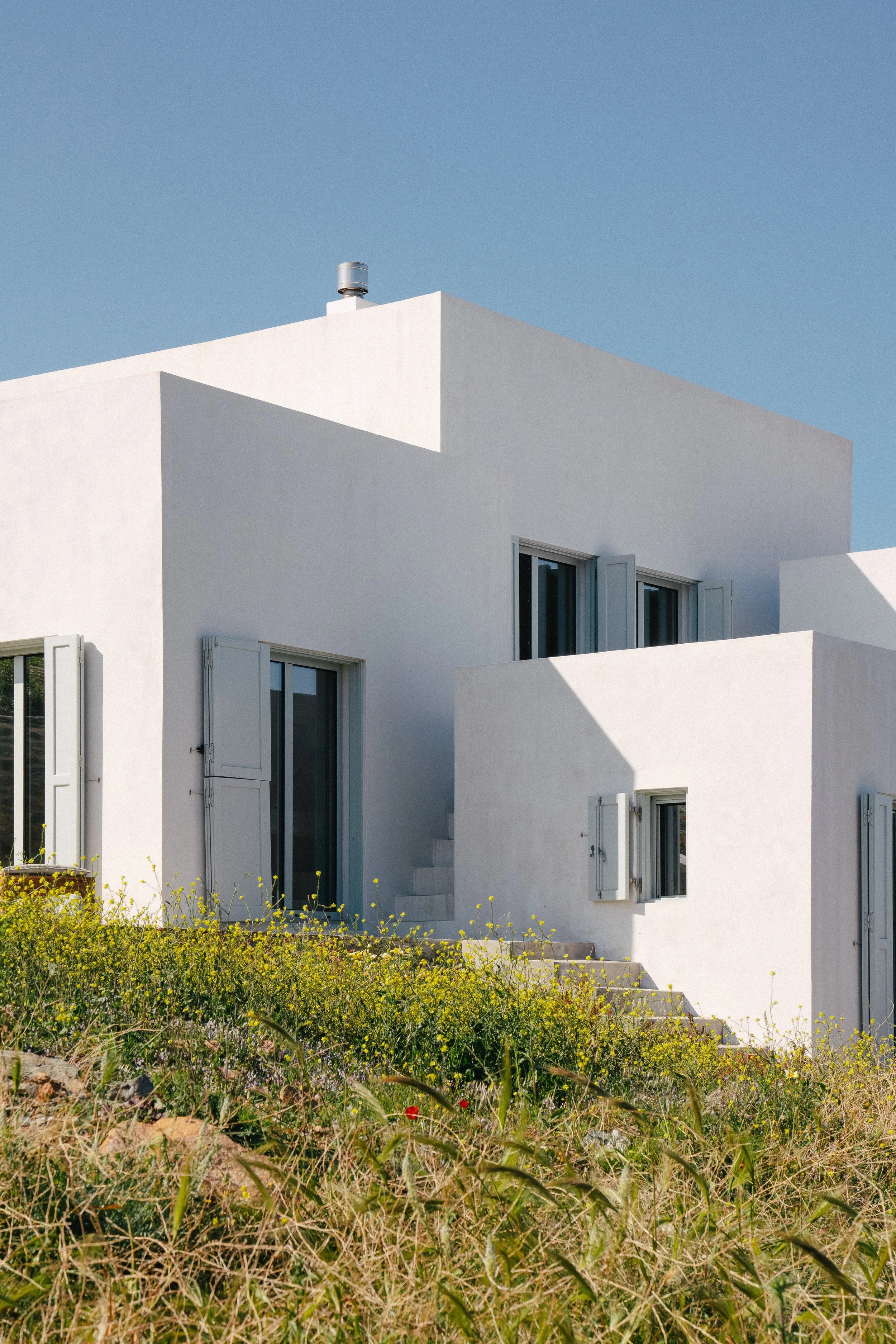A white modern house with multiple levels, open windows, and a clear blue sky, surrounded by green grass and wildflowers.