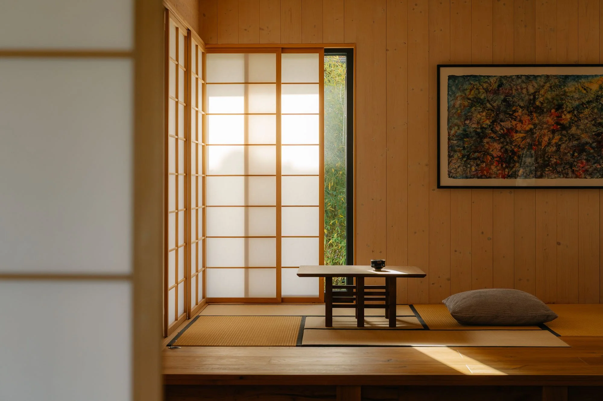 A minimalist Japanese-style room with wooden walls and a sliding shoji door, a low table with a small black bowl, a gray floor pillow, and a large colorful framed artwork on the wall.