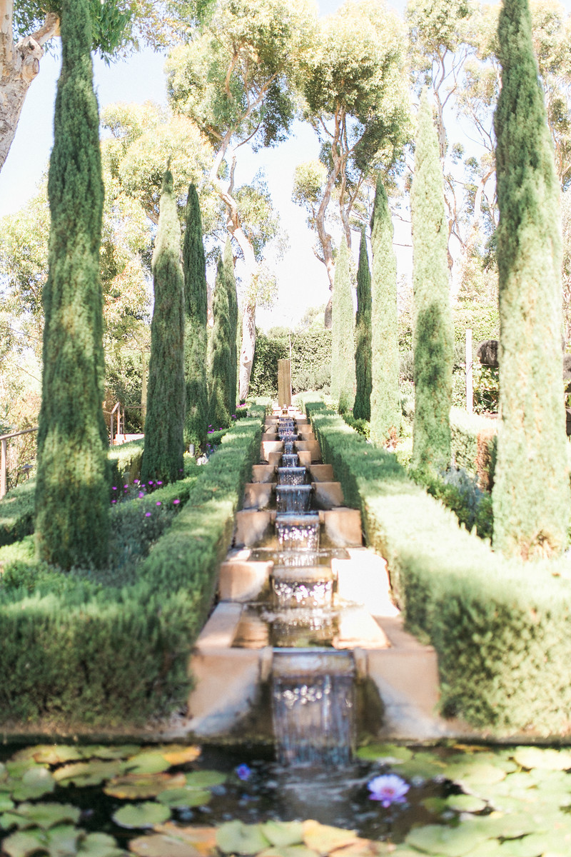 A peaceful garden with a small cascading water fountain, tall cypress trees lining both sides, and a pond with lily pads and pink water lilies in the foreground.