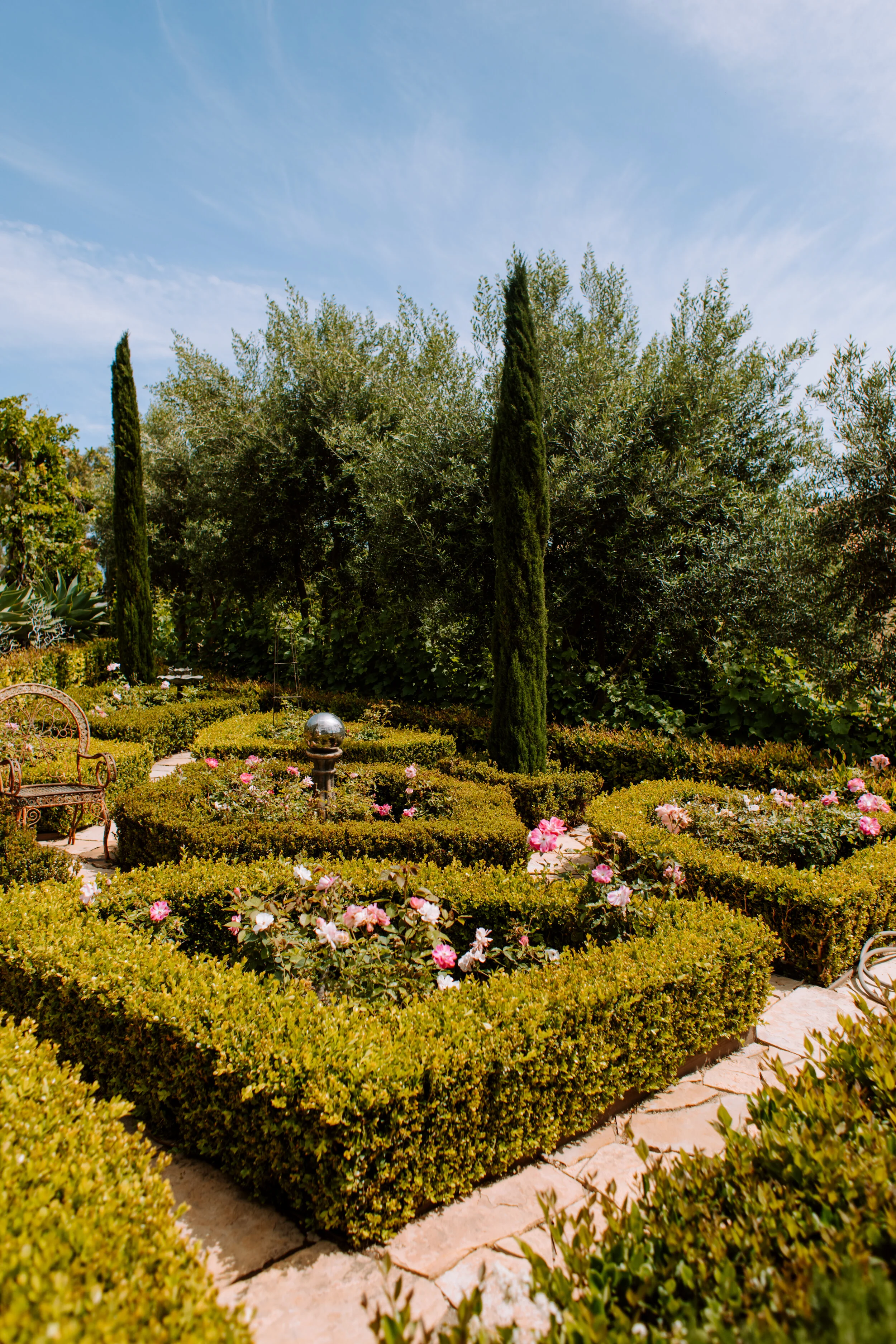 A well-maintained garden with neatly trimmed bushes, pink and white flowers, tall cypress trees, and a rustic metal bench under a bright blue sky.